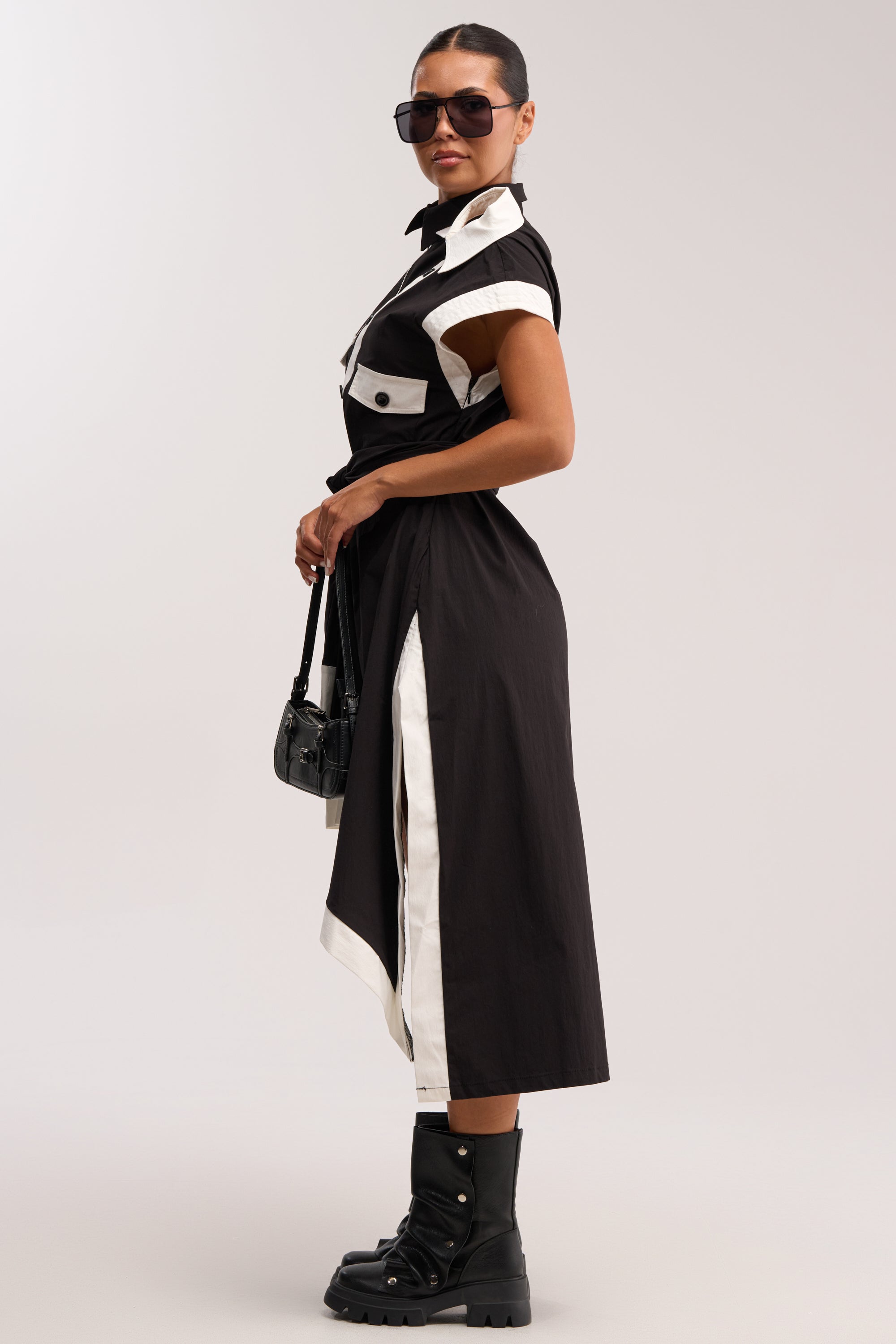A woman wearing sunglasses and the NO FILTER NEEDED BUTTON UP MAXI DRESS with side slits, styled with a small black handbag and chunky boots, stands sideways against a plain background.