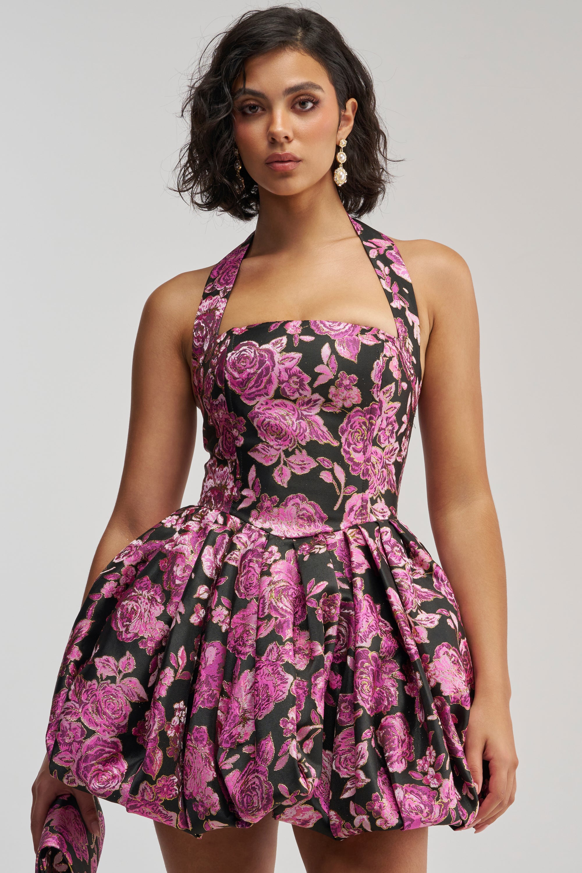 Wearing the CATCH ME DANCING FLORAL MINI DRESS—a black halter-neck mini dress with a full, bubble skirt and vibrant pink floral pattern—she accessorizes with a matching clutch, gold earrings, and styles her short, wavy dark hair for a striking look.
