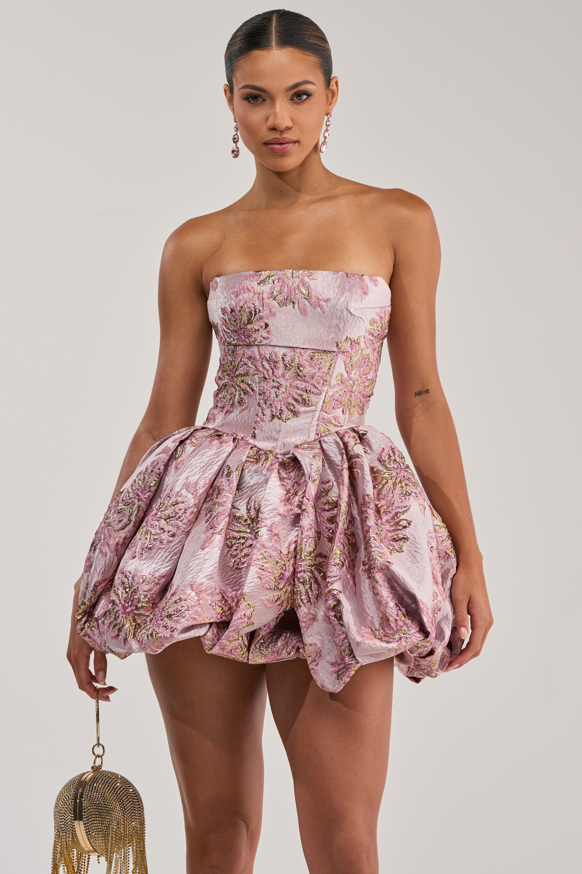 A woman stands in the GLAM GROOVE BROCADE MINI DRESS, featuring a strapless pink floral brocade design with a voluminous bubble skirt. She pairs it with drop earrings and a gold fringe bag for a chic homecoming look.