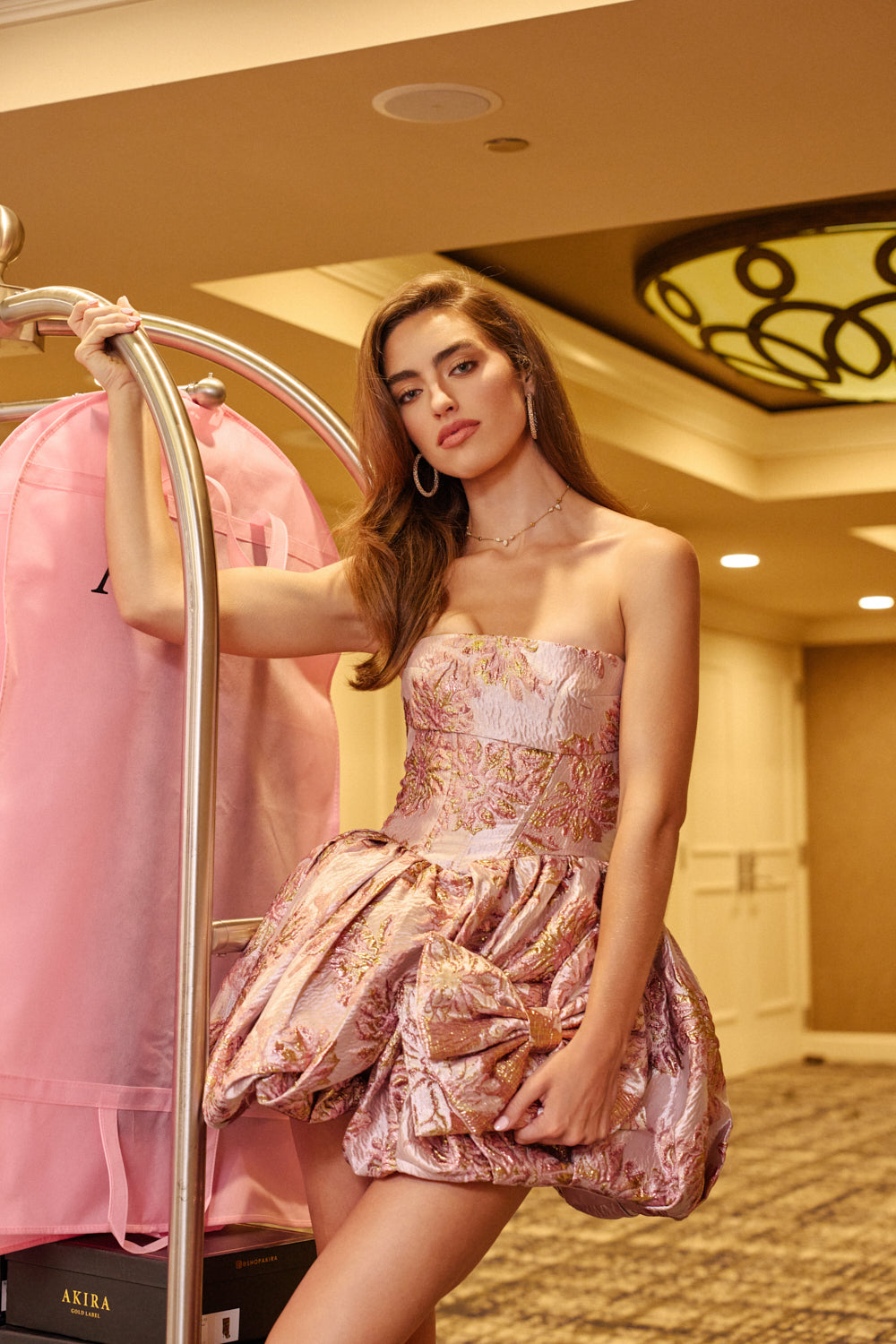 Wearing the GLAM GROOVE BROCADE MINI DRESS, a woman with long hair and hoop earrings poses indoors beside a garment bag on a luggage cart. The strapless mini dress features a pink, floral-patterned brocade design, making it an elegant choice for any special occasion. She stands in a well-lit hallway with stylish decor, completing the sophisticated look.