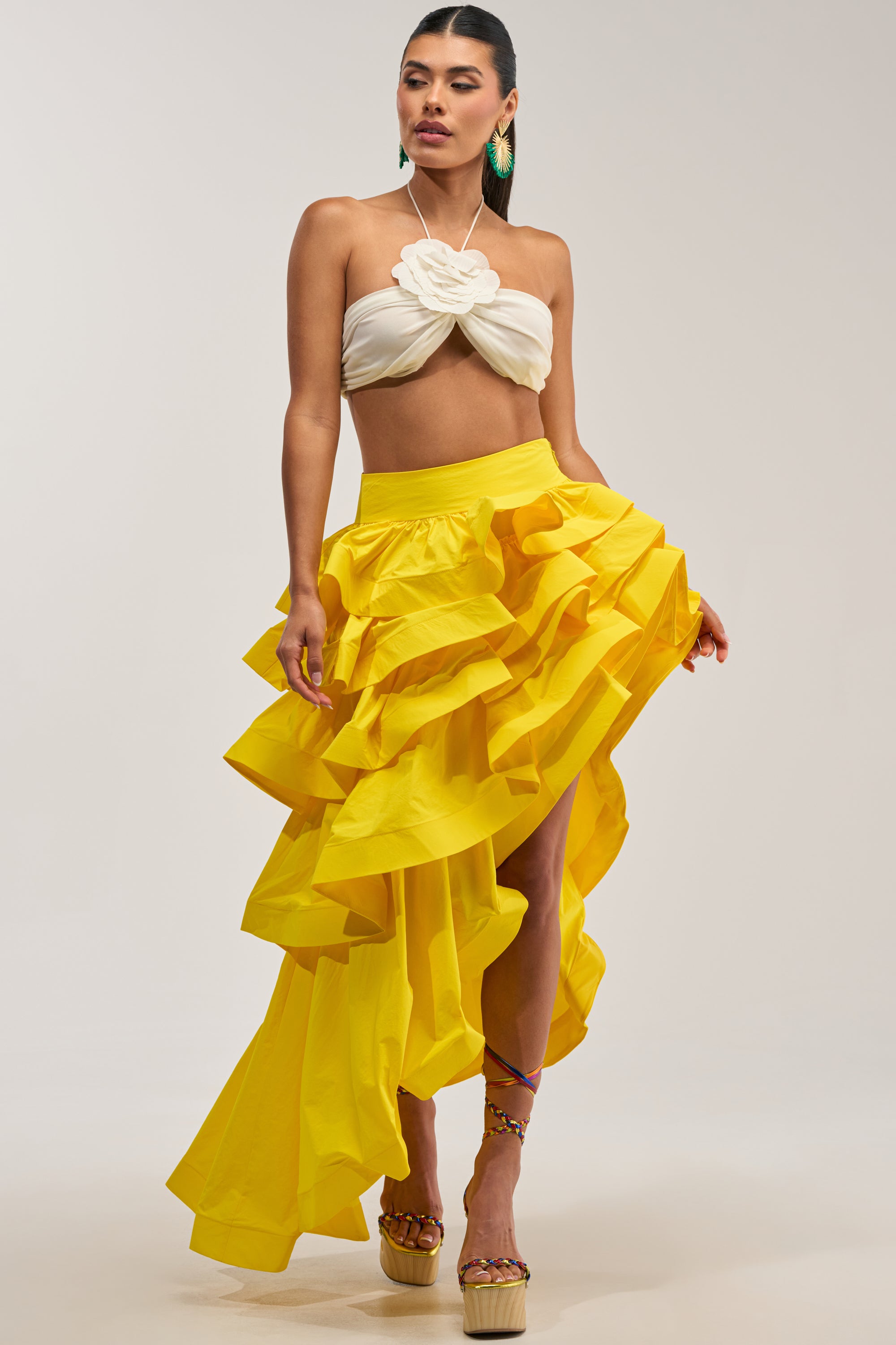 A model showcases the ELEGANT QUEEN ASYMMETRICAL SKIRT IN YELLOW, styled with a white crop top featuring bold floral detailing. The skirt’s striking high-low ruffle hem creates an eye-catching silhouette, complemented by statement green leaf earrings and platform wedges with colorful braided straps—an ideal ensemble for stylish occasion dressing against a solid light gray background.