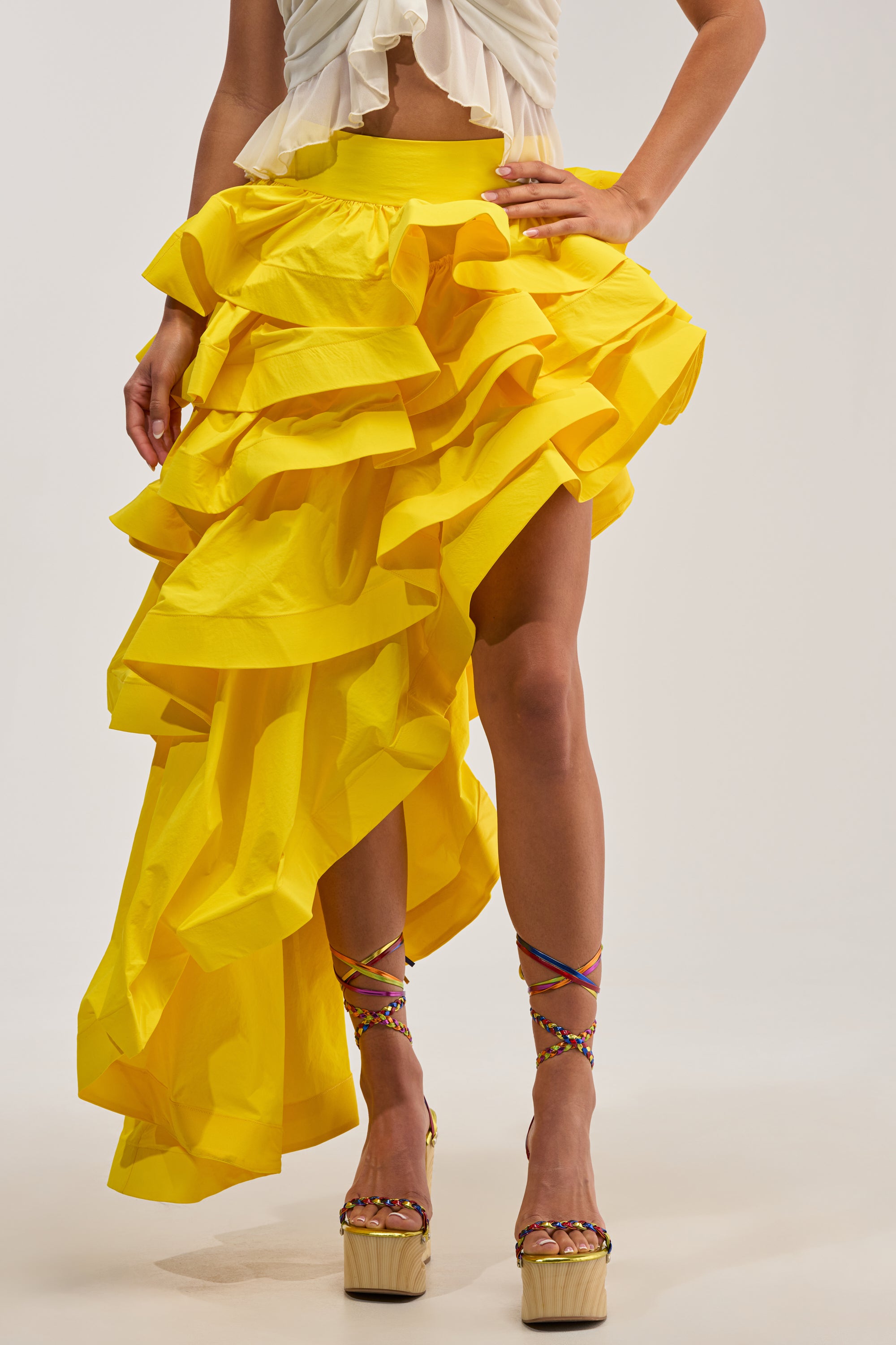 A person wearing the ELEGANT QUEEN ASYMMETRICAL SKIRT IN YELLOW, featuring a striking ruffle design, pairs it with strappy platform heels while posing against a plain background, perfectly embodying chic occasion dressing.