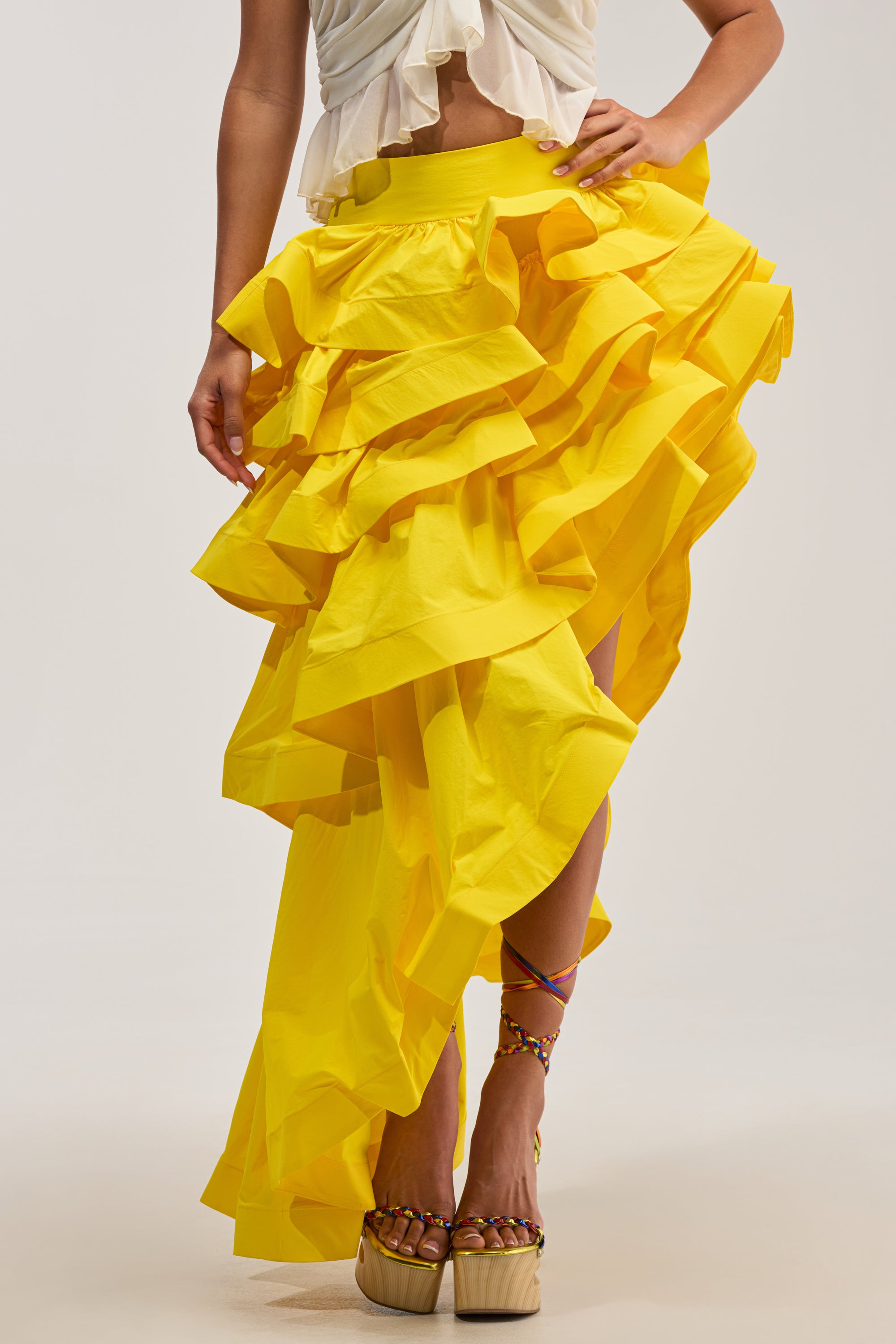 Wearing the ELEGANT QUEEN ASYMMETRICAL SKIRT IN YELLOW, this person pairs its vibrant ruffles and high-low, asymmetrical hem with a white ruffled top. Colorful thin-strapped sandals with wooden platform soles complete the effortlessly chic look—perfect for occasion dressing. The backdrop is a simple, neutral studio setting.