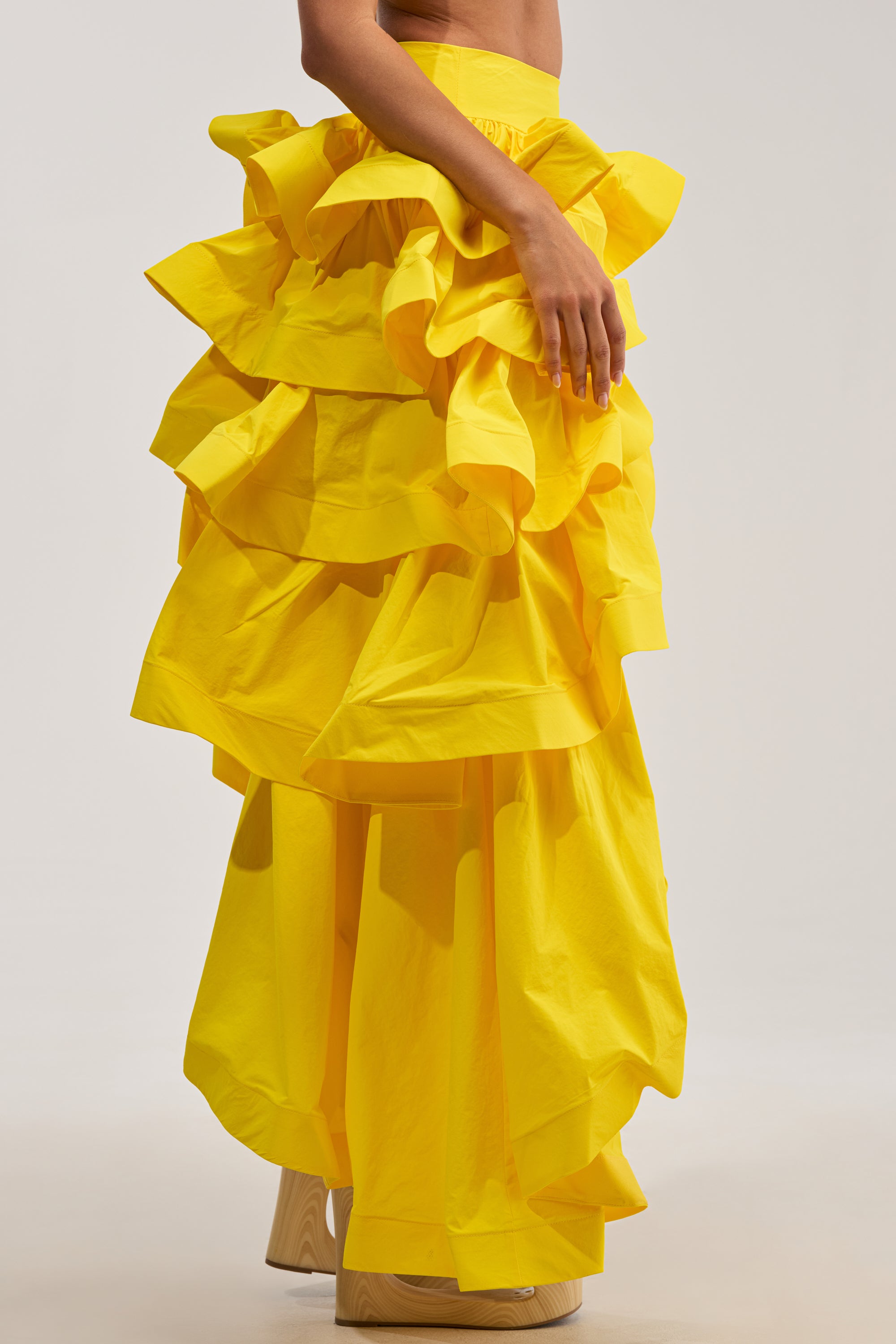 A person models the ELEGANT QUEEN ASYMMETRICAL SKIRT IN YELLOW against a plain backdrop, showing only the lower torso and upper legs. One hand rests on the hip, highlighting the skirt's bold, asymmetrical silhouette with its dramatic layers—a standout choice for special occasions.