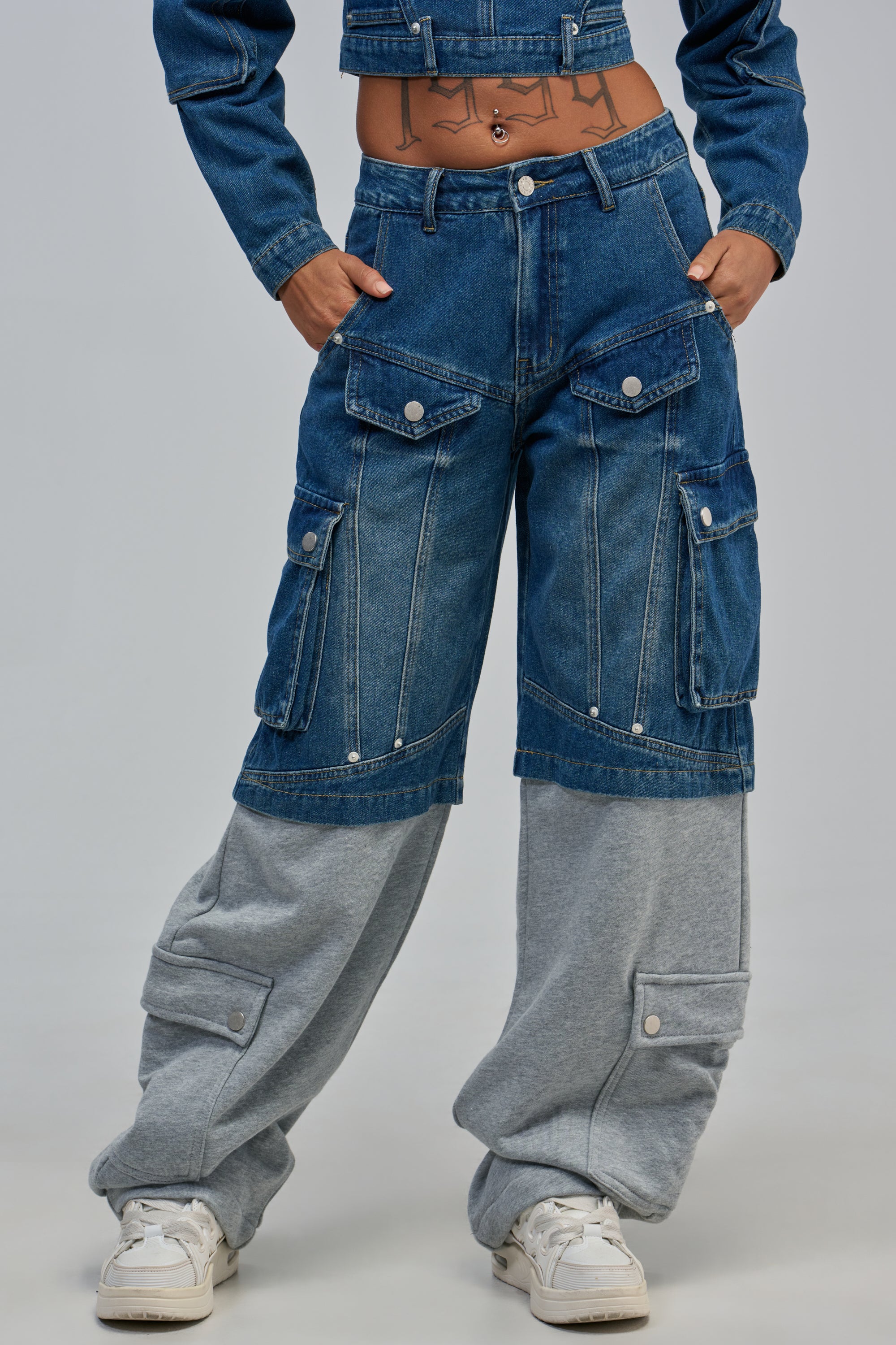A person wears THE IT GIRL DENIM PANT with a cropped denim jacket and white sneakers. Their hands rest on their hips, revealing a tattoo on their exposed midriff.