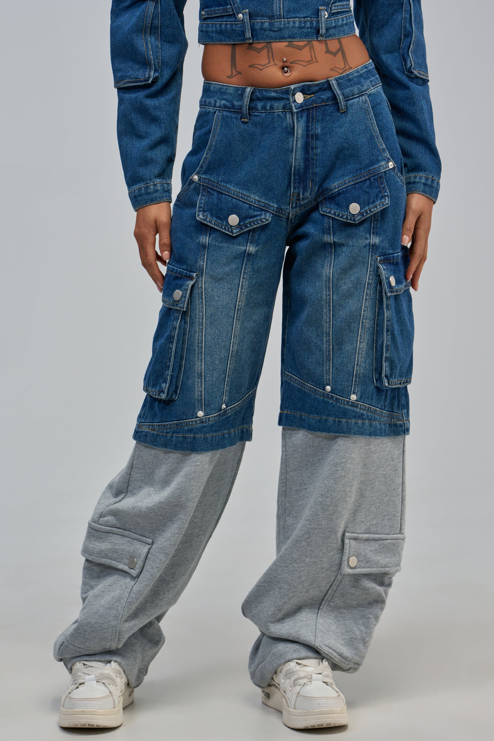 Woman wearing THE IT GIRL DENIM PANT with split denim, gray sweatpant knees, cargo pockets, paired with a cropped denim jacket and white sneakers. Only her torso and legs show against a plain background.
