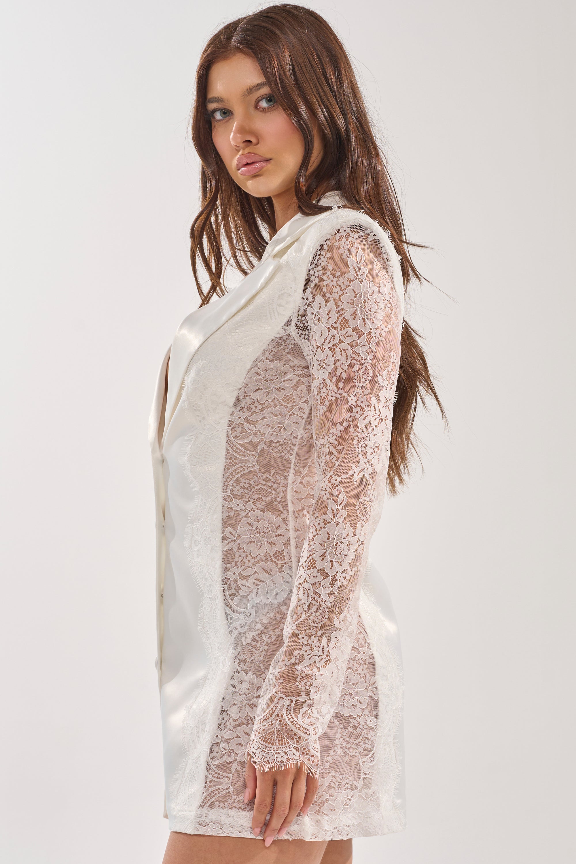 A woman models the SHE MEANS BUSINESS BLAZER MINI DRESS IN WHITE, featuring sheer floral lace sleeves and lace side panels, standing against a plain light background and looking directly at the camera. 2