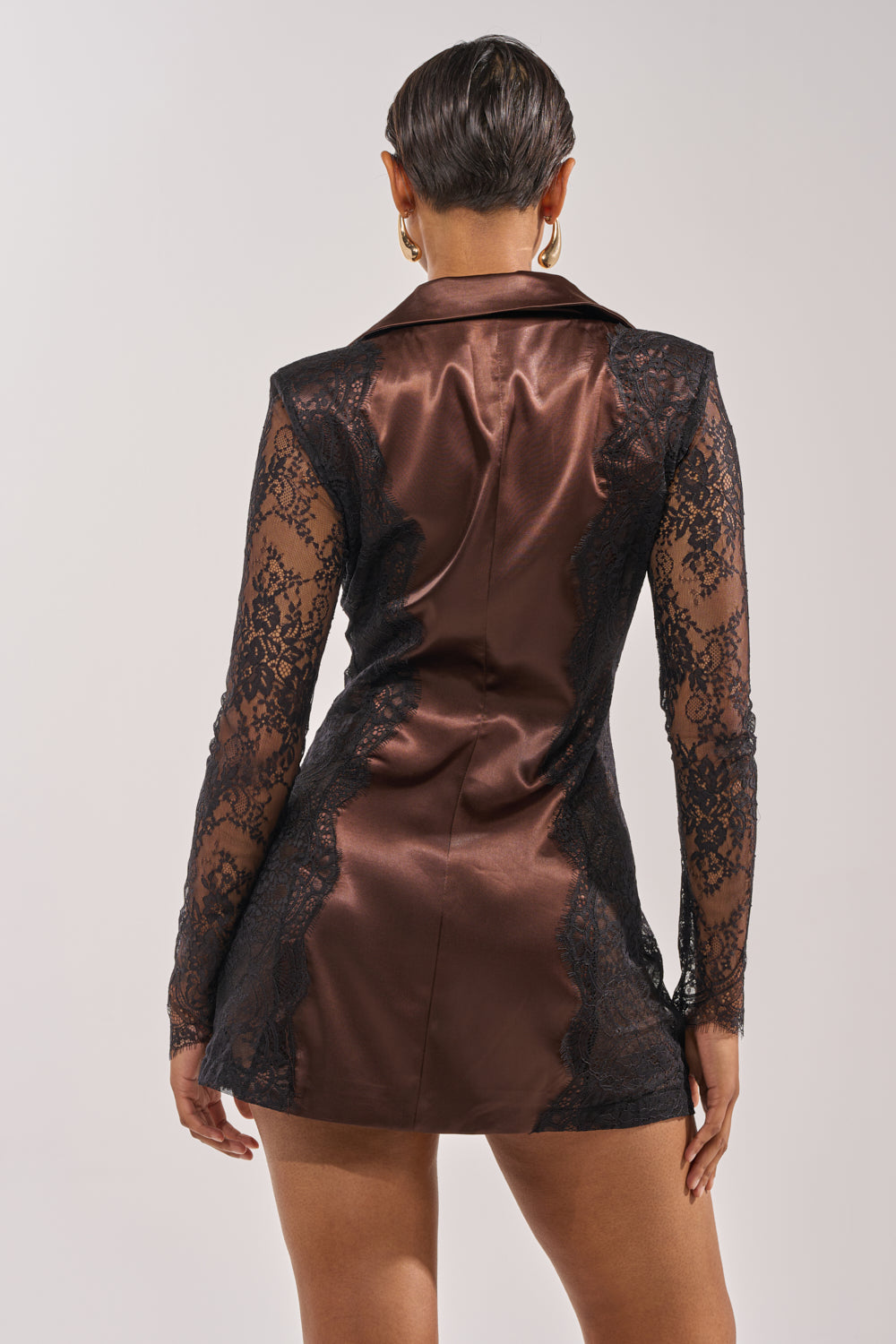 A woman is seen from the back wearing the SHE MEANS BUSINESS BLAZER MINI DRESS—brown satin with black lace long sleeves and side accents. Her hair is slicked back, paired with gold hoop earrings. 3