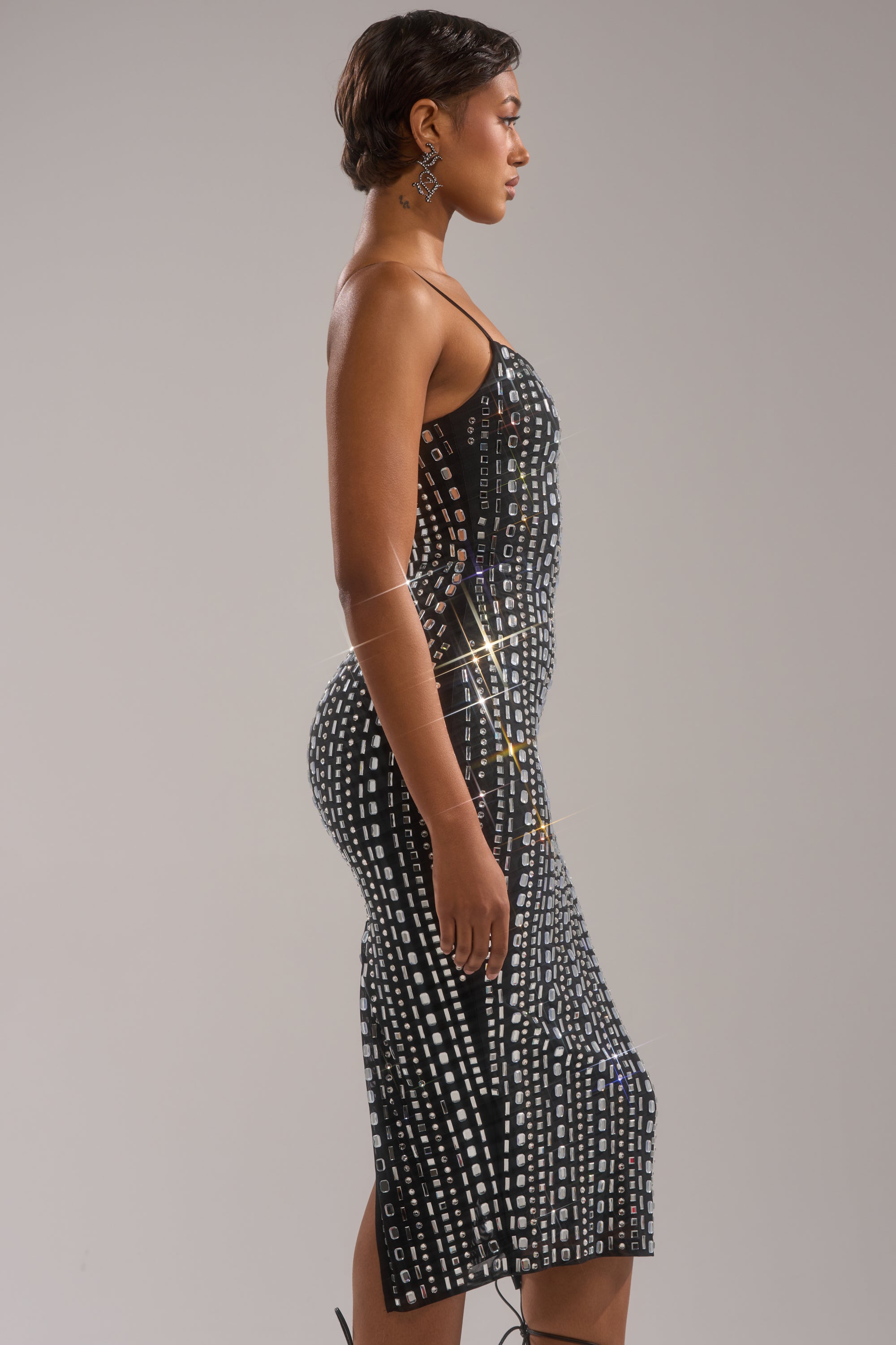 A woman in profile wears the GIA RHINESTONE MAXI DRESS—a fitted, sleeveless dress adorned with geometric rhinestones and silver sequins—paired with geometric earrings and sporting short hair, set against a plain gray background. 2