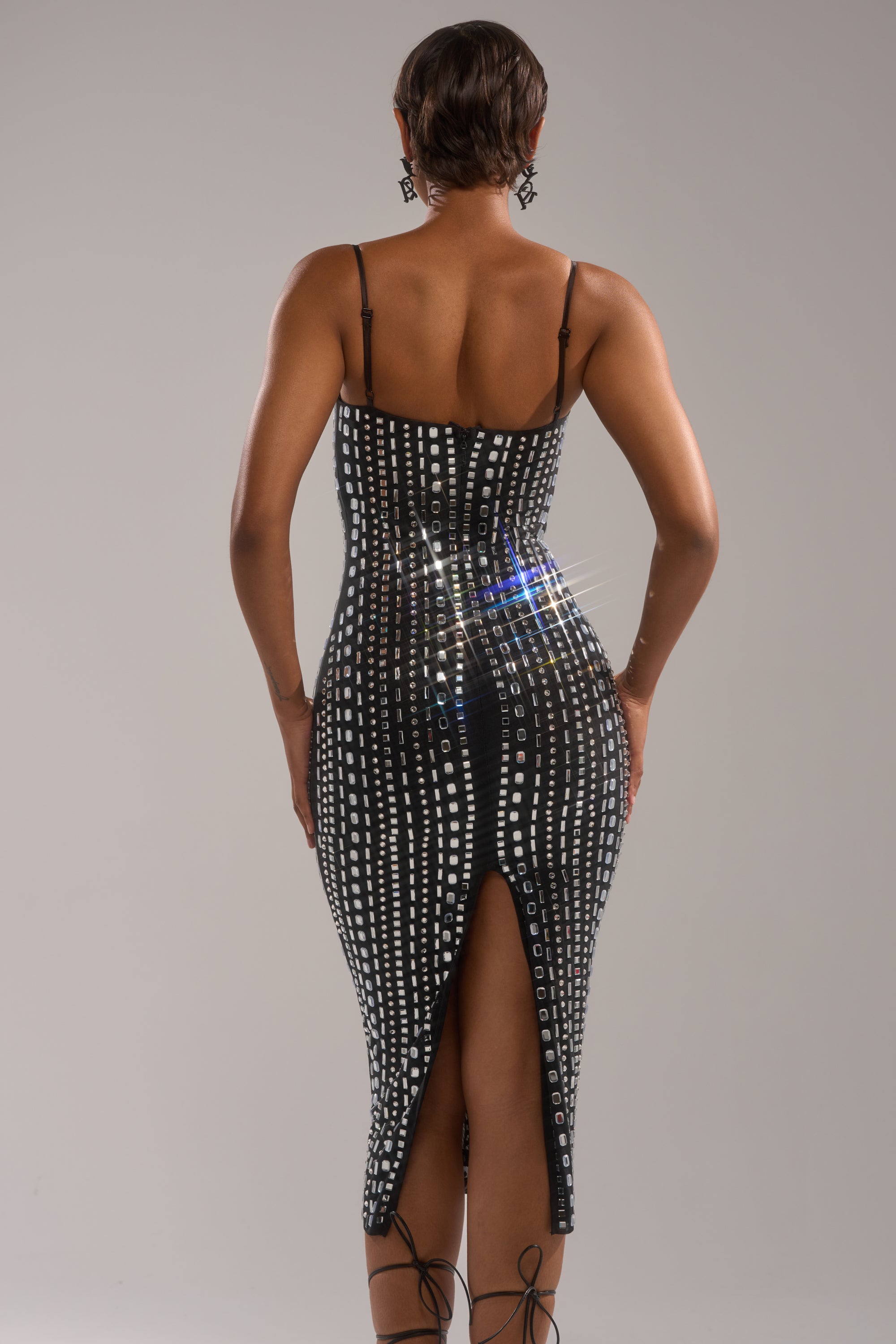 A person stands facing away, wearing the GIA RHINESTONE MAXI DRESS—a fitted black maxi with white square rhinestone patterns, thin straps, and a back slit. Styled with black high-heeled sandals and earrings for an elegant look. 3