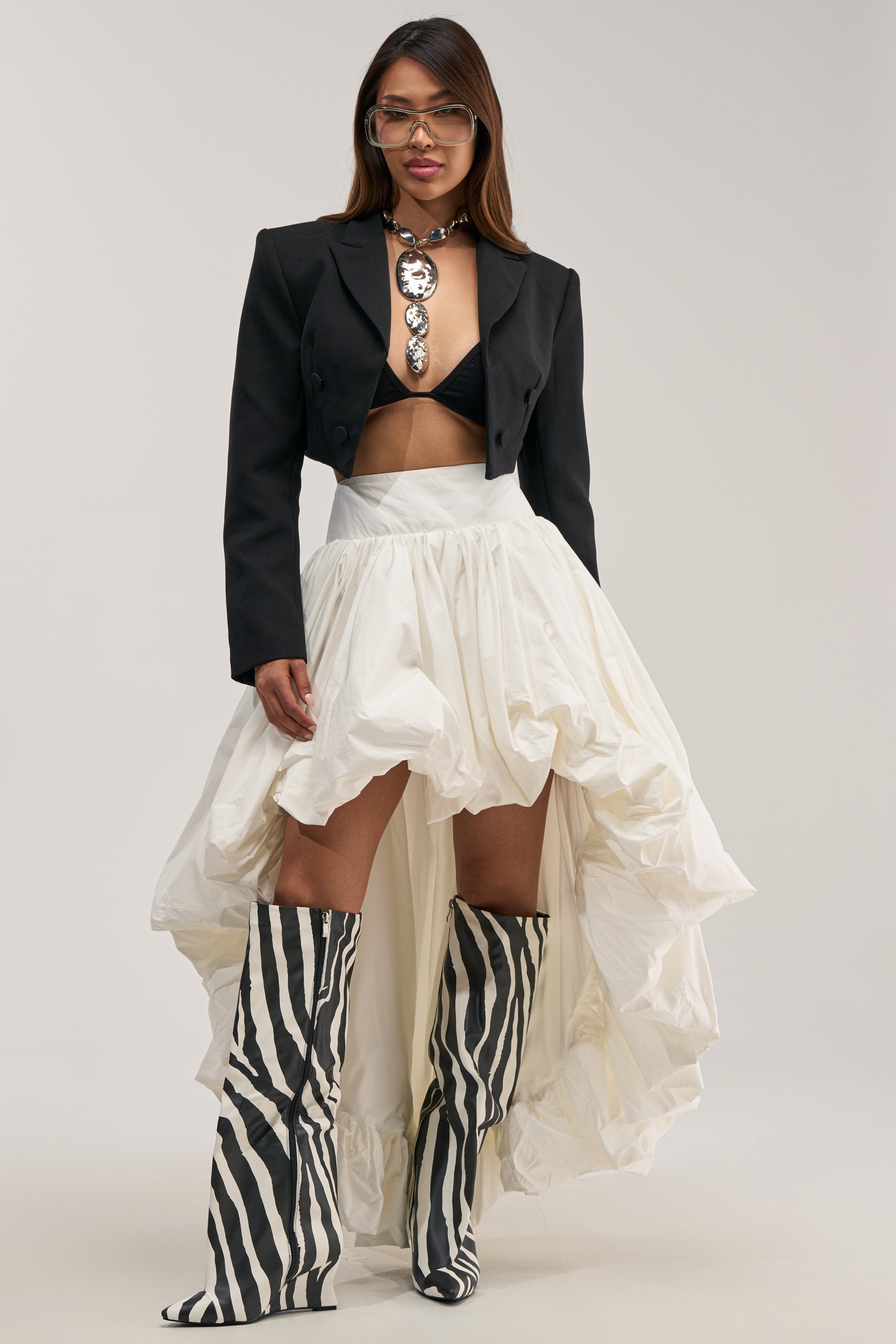 A woman stands confidently against a plain background, styled in a black cropped blazer, black bra, statement necklace, and THE BIGGER THE BETTER HIGH LOW MAXI SKIRT—a couture-inspired white ruffled high-low piece—paired with knee-high zebra-print boots.