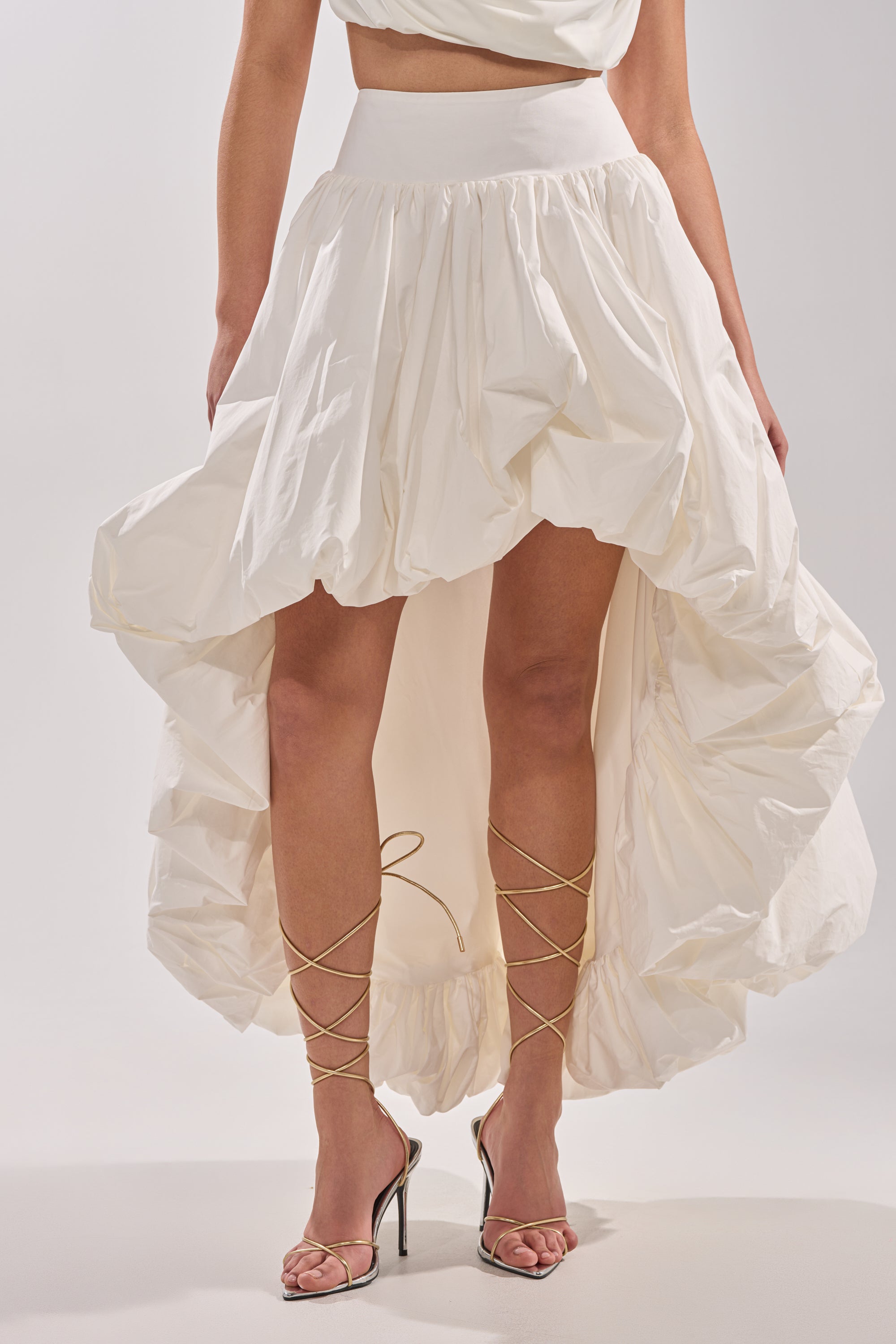 Someone wears THE BIGGER THE BETTER HIGH LOW MAXI SKIRT, featuring a voluminous ruffled hem, styled with strappy high-heeled sandals. The image highlights the lower half against a plain background.