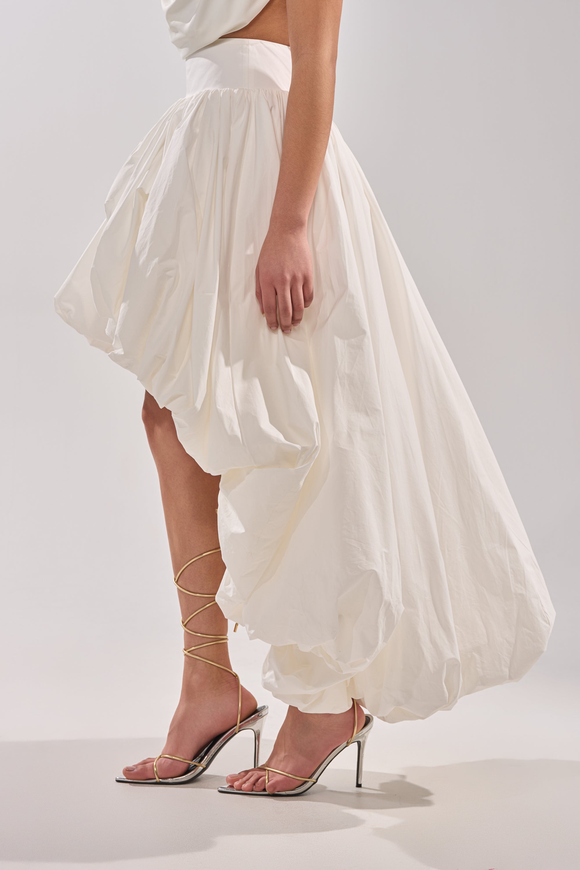 A person wearing THE BIGGER THE BETTER HIGH LOW MAXI SKIRT in white and strappy metallic heels stands against a plain light background. Only their lower half is visible, emphasizing the skirt's voluminous silhouette.