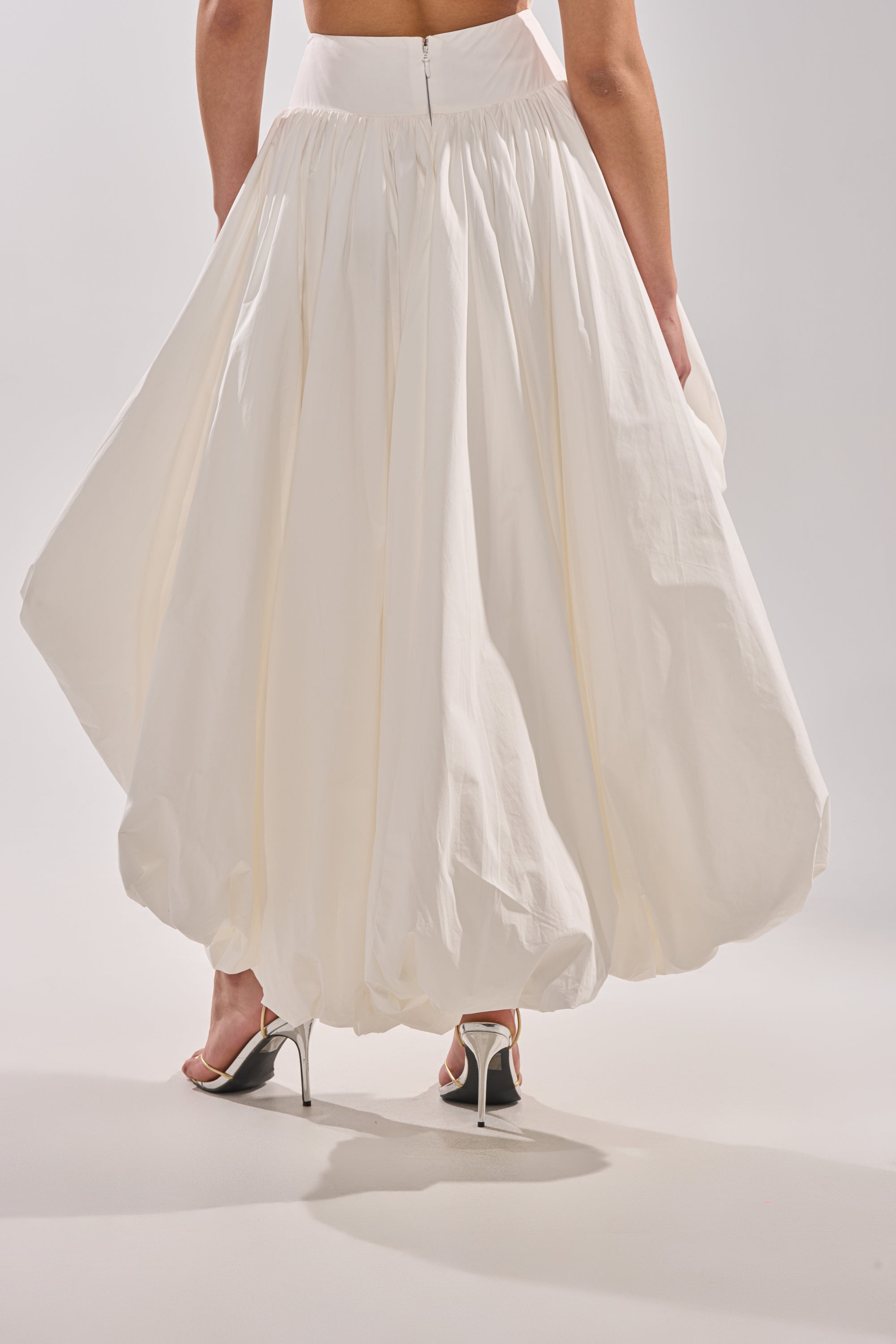 Someone wearing THE BIGGER THE BETTER HIGH LOW MAXI SKIRT, a voluminous, ankle-length piece with a gathered hem, paired with silver high-heeled sandals and photographed from behind against a plain background.