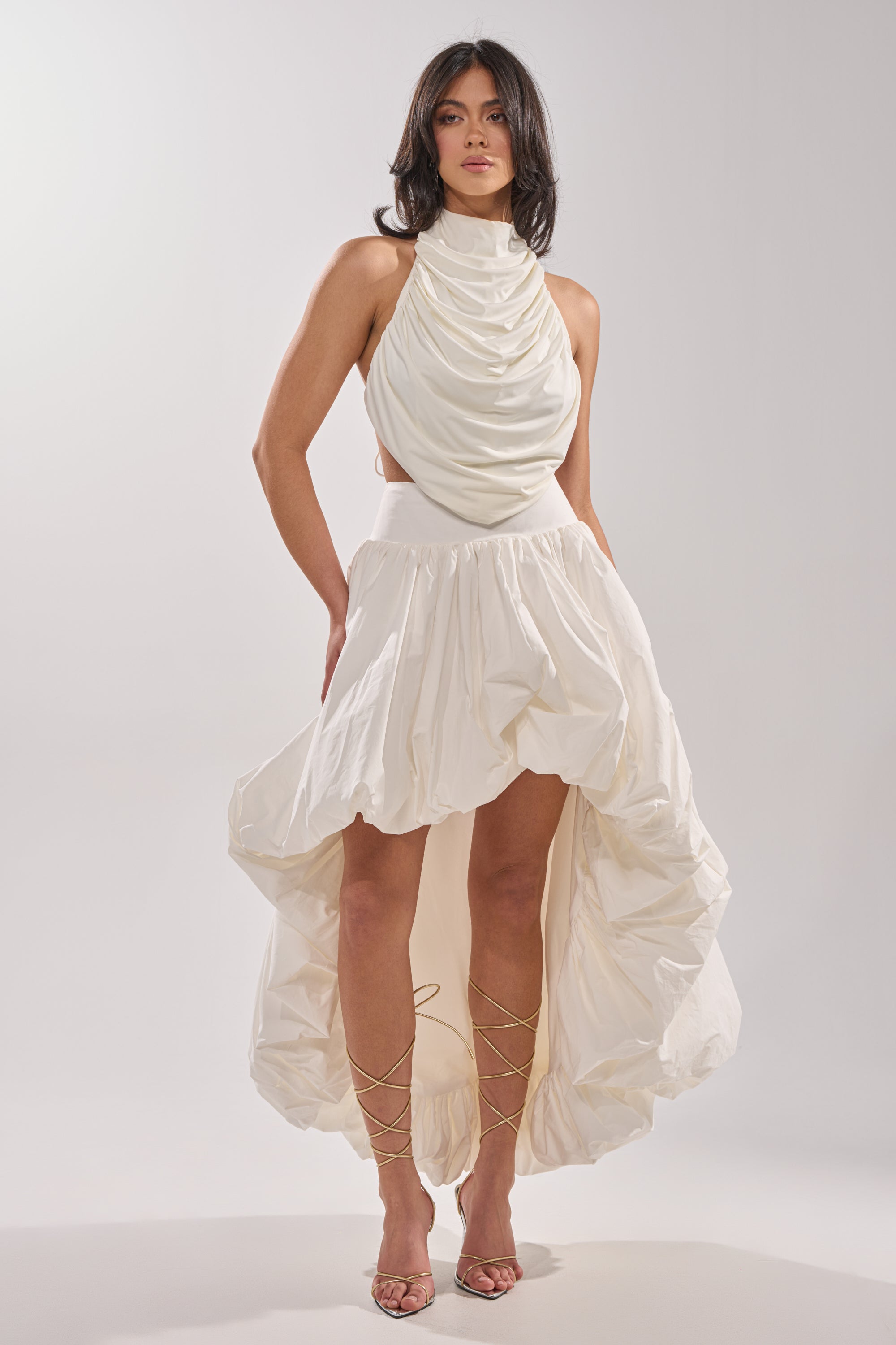 A woman stands against a plain background wearing a sleeveless white dress with a draped halter neckline and THE BIGGER THE BETTER HIGH LOW MAXI SKIRT, paired with strappy heeled sandals.