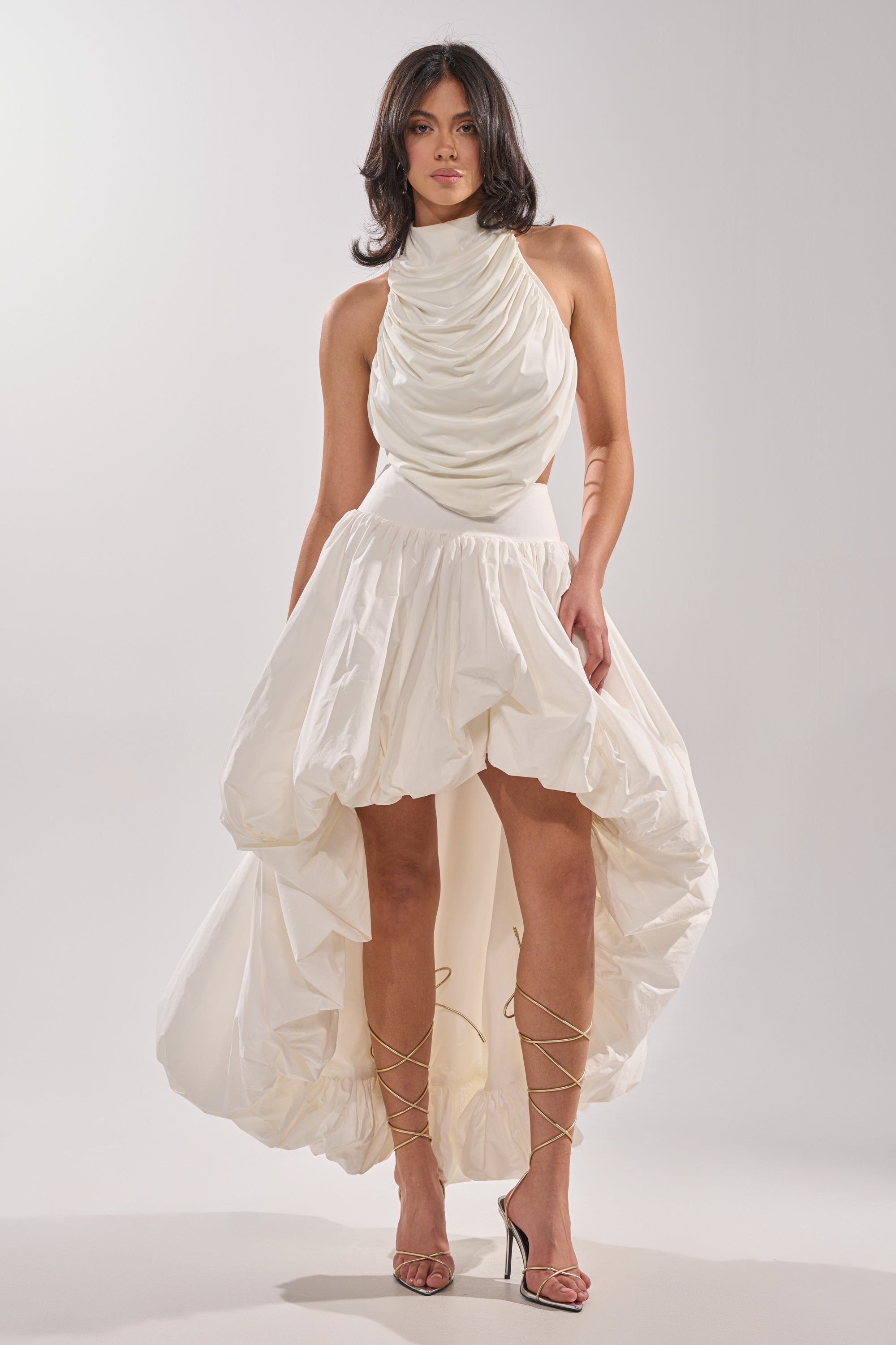 A woman wears THE BIGGER THE BETTER HIGH LOW MAXI SKIRT, featuring a statement high-low hem and draped halter neckline, paired with strappy high-heeled sandals. She stands against a plain, light-colored background.