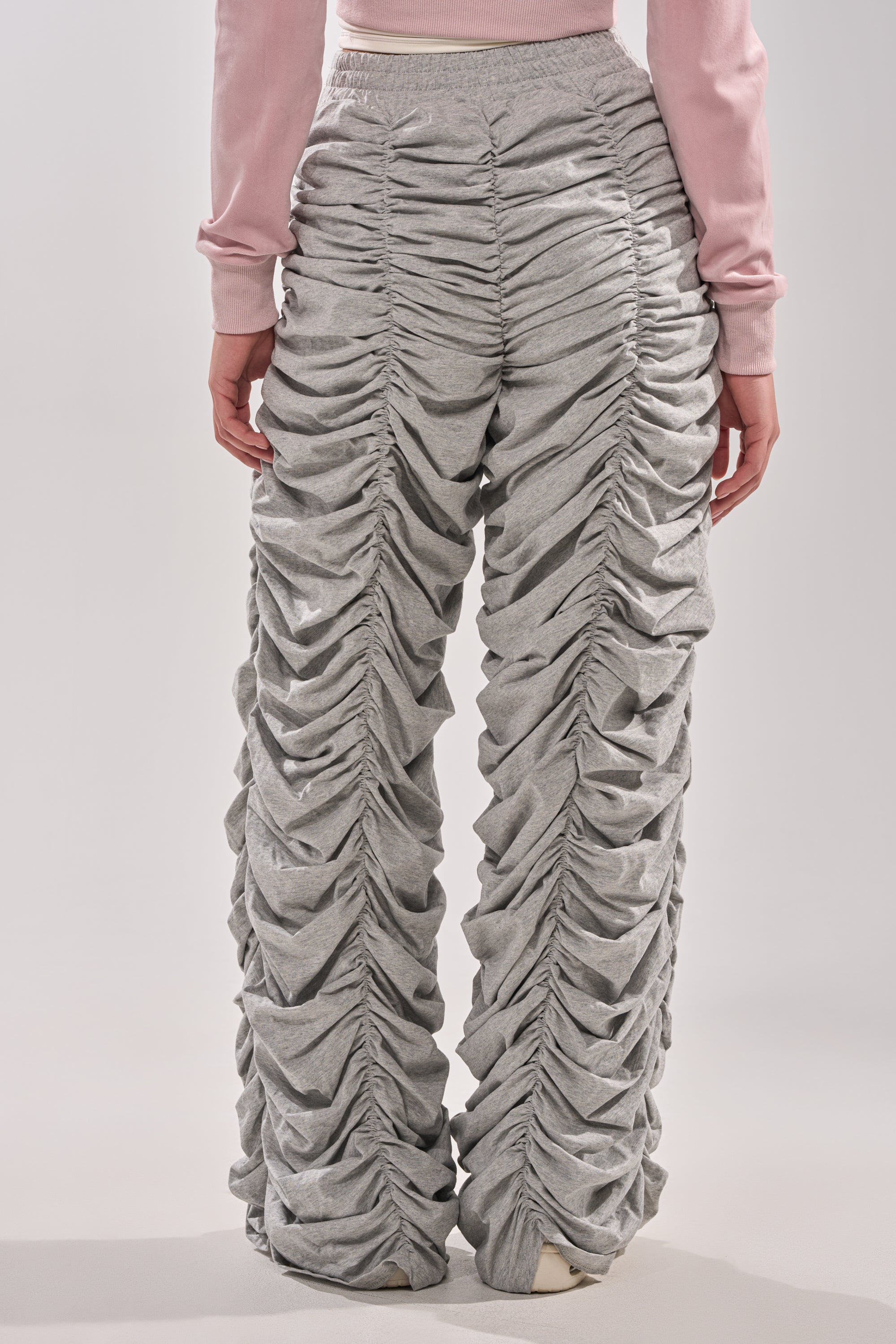 A person is seen from the back wearing AKIRA Label’s IM THAT TYPE RUCHED JOGGER in heather grey, featuring a high waist and ruffled, gathered texture down the legs, paired with a pink long-sleeve top. 3