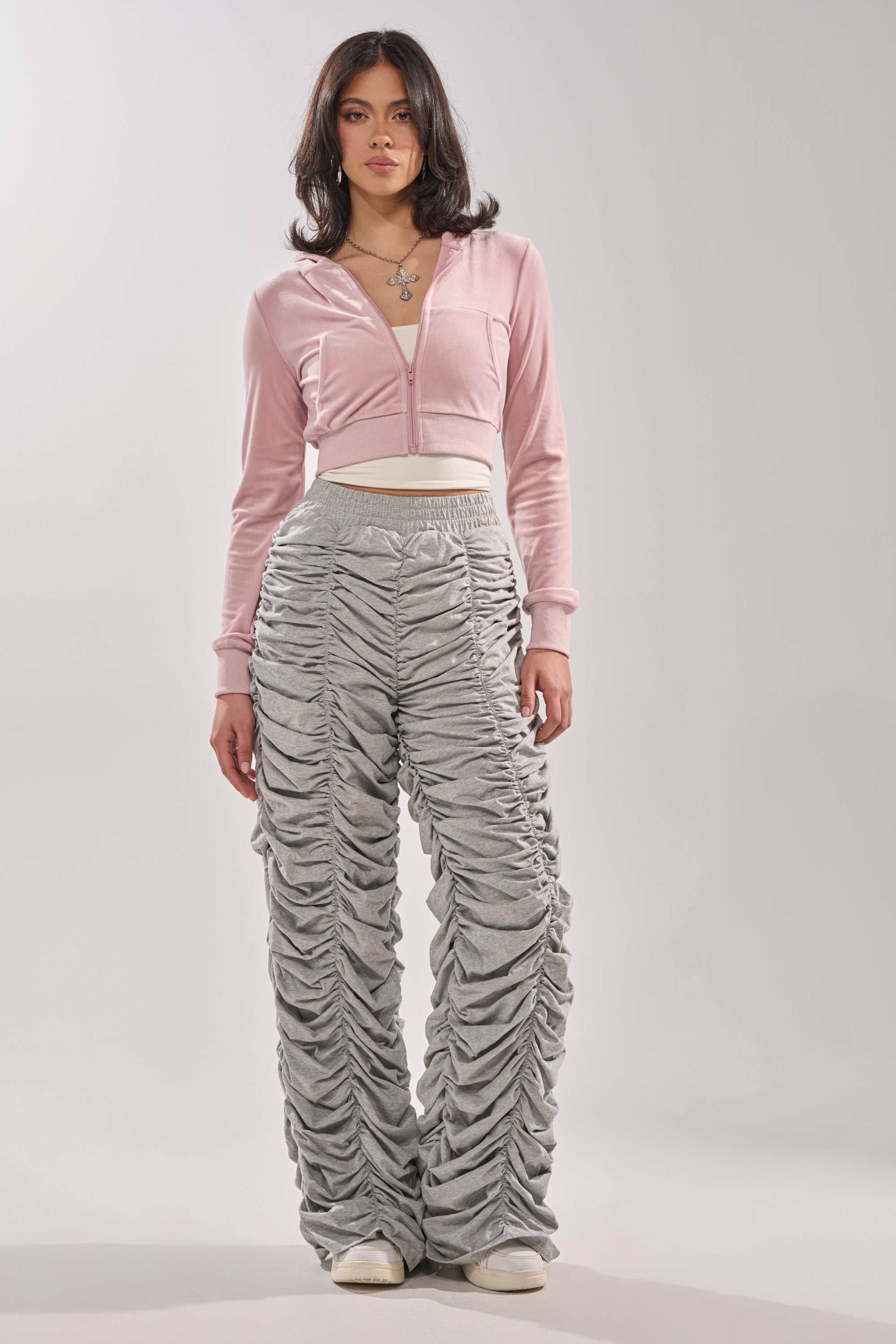 A woman with shoulder-length dark hair poses in a relaxed stance against a plain background, wearing an AKIRA Label light pink cropped zip-up hoodie, IM THAT TYPE RUCHED JOGGER heather grey joggers, white sneakers, and layered necklaces. 4