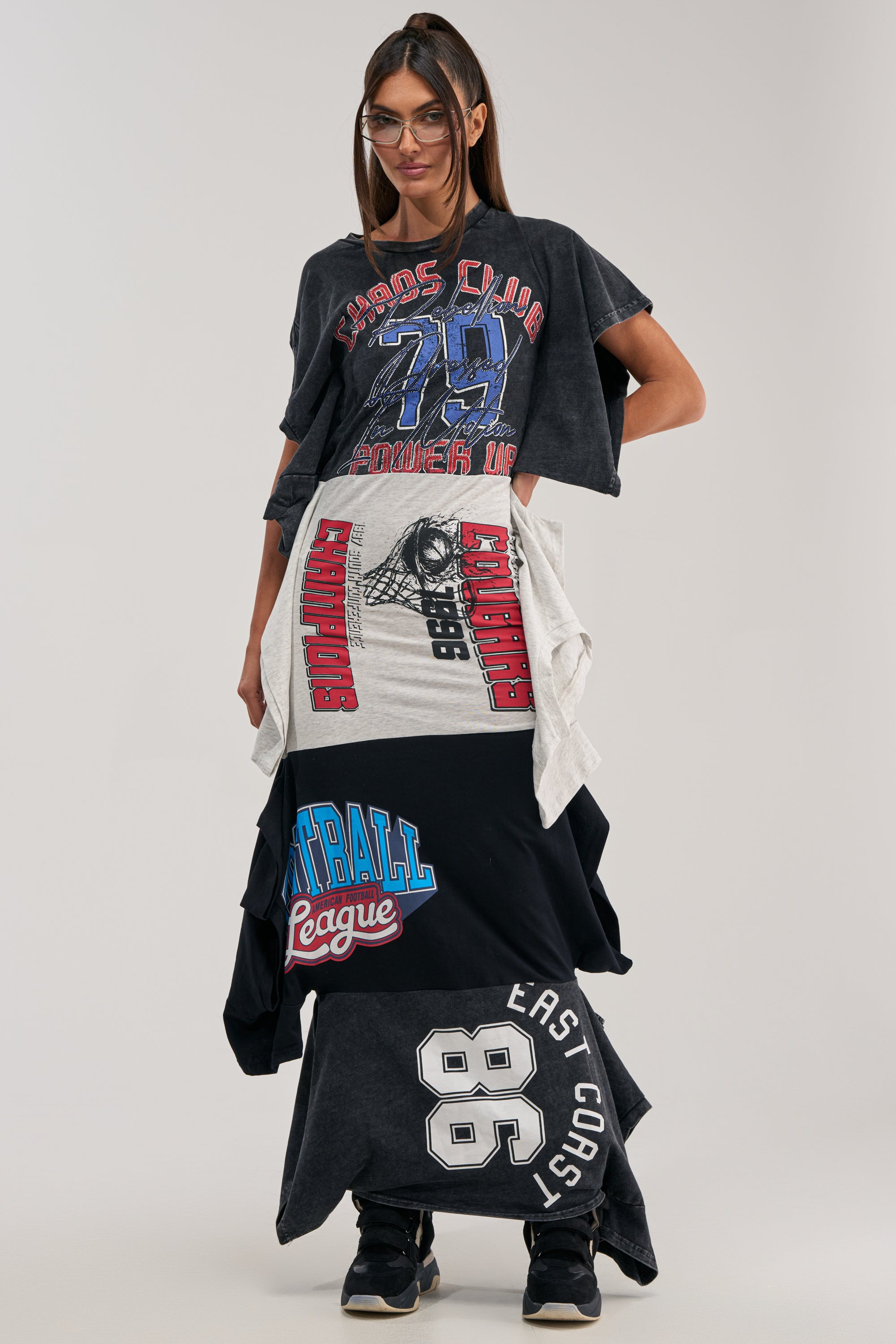 A person with long hair and glasses stands confidently against a plain light background, styled in streetwear fashion wearing the YOUR BIGGEST FAN GRAPHIC TEE MAXI DRESS with layered, oversized skirts.