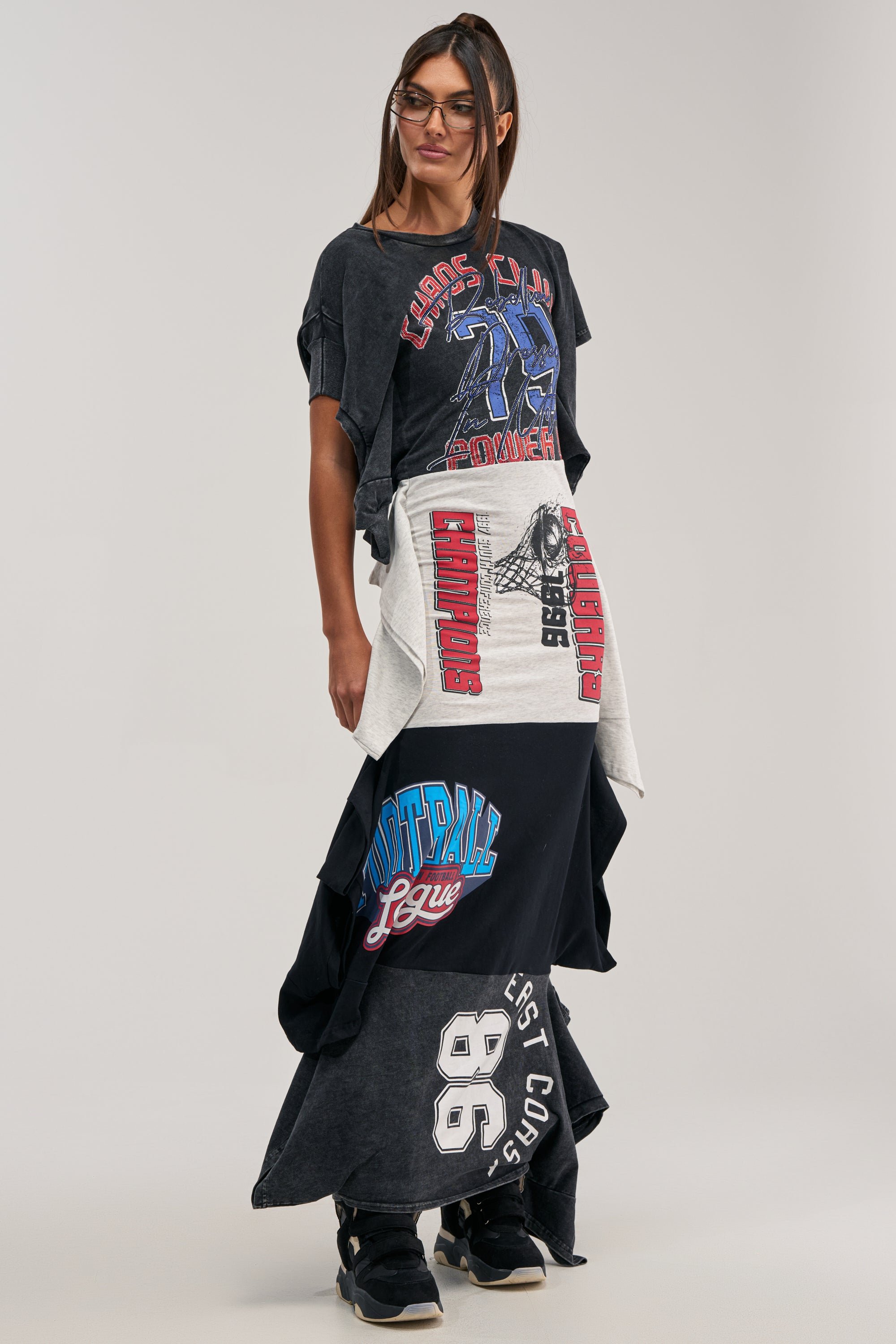 A woman with long hair and glasses styles the YOUR BIGGEST FAN GRAPHIC TEE MAXI DRESS with black sneakers, creating an effortlessly cool streetwear look against a plain background.