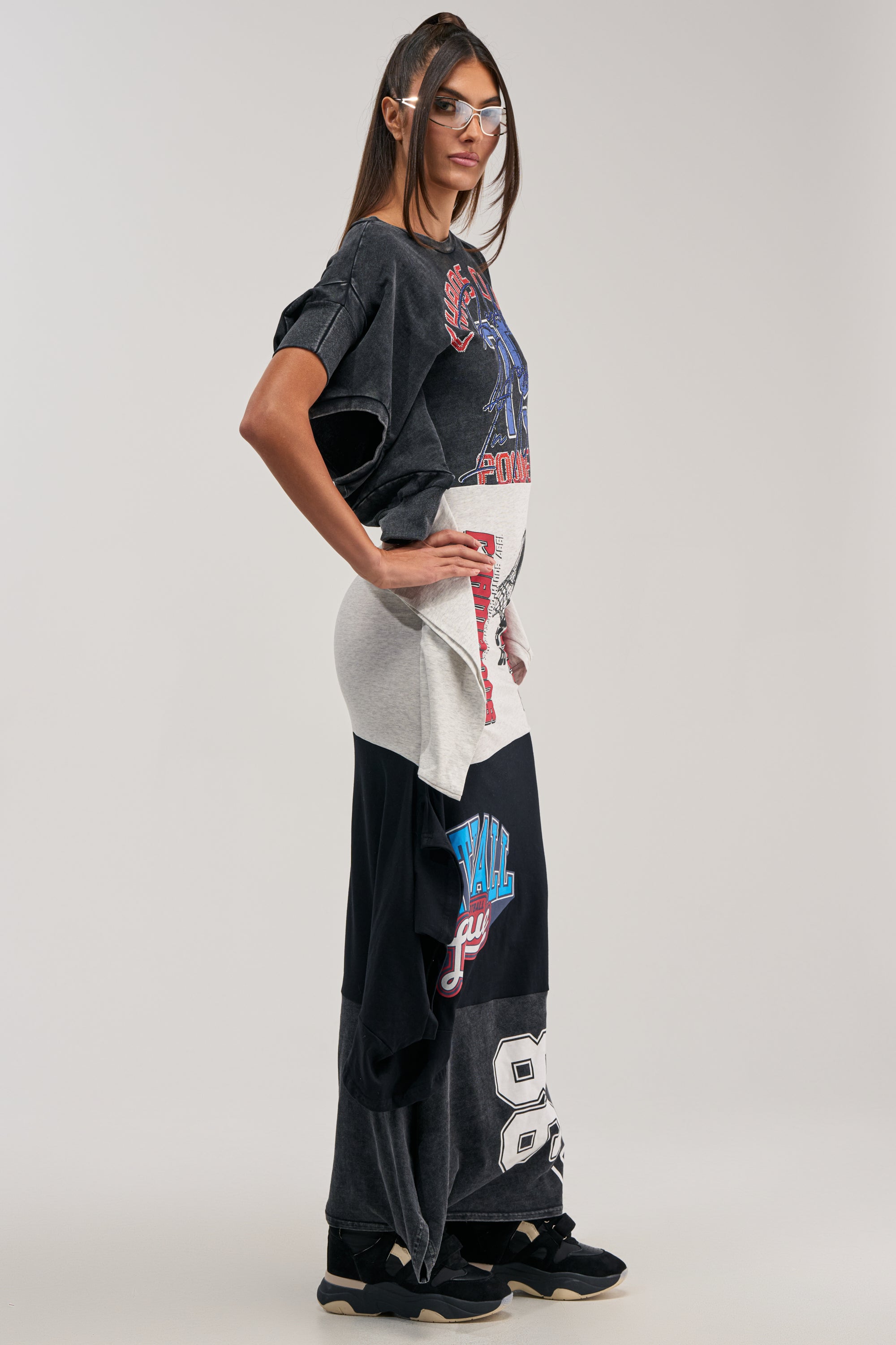 A woman in sporty, layered streetwear stands sideways in a plain light gray setting. She wears the YOUR BIGGEST FAN GRAPHIC TEE MAXI DRESS with patchwork sports numbers and logos, black sneakers, and clear safety glasses.