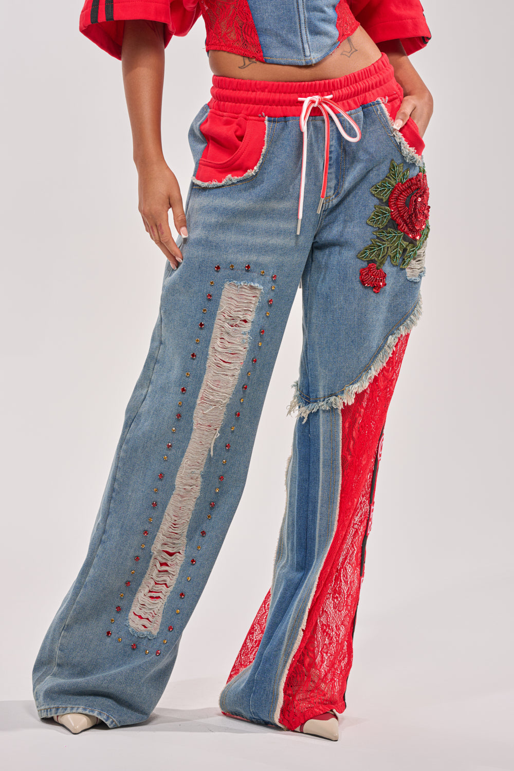 Someone rocks the GAME TIME JOGGER—wide-leg patchwork denim joggers with red lace panels, distressed details, stud accents, and an embroidered red rose—paired with a cropped red and blue top for standout streetwear. 3