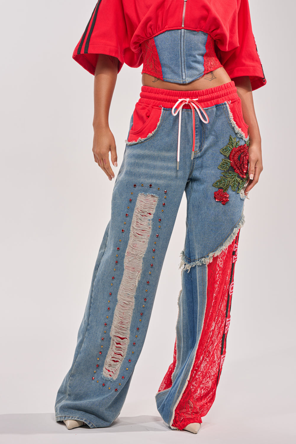 A person sports the GAME TIME JOGGER: patchwork denim joggers with distressed touches, red laces, floral embroidery, paired with a matching red top featuring a denim corset and drawstring waist. 0