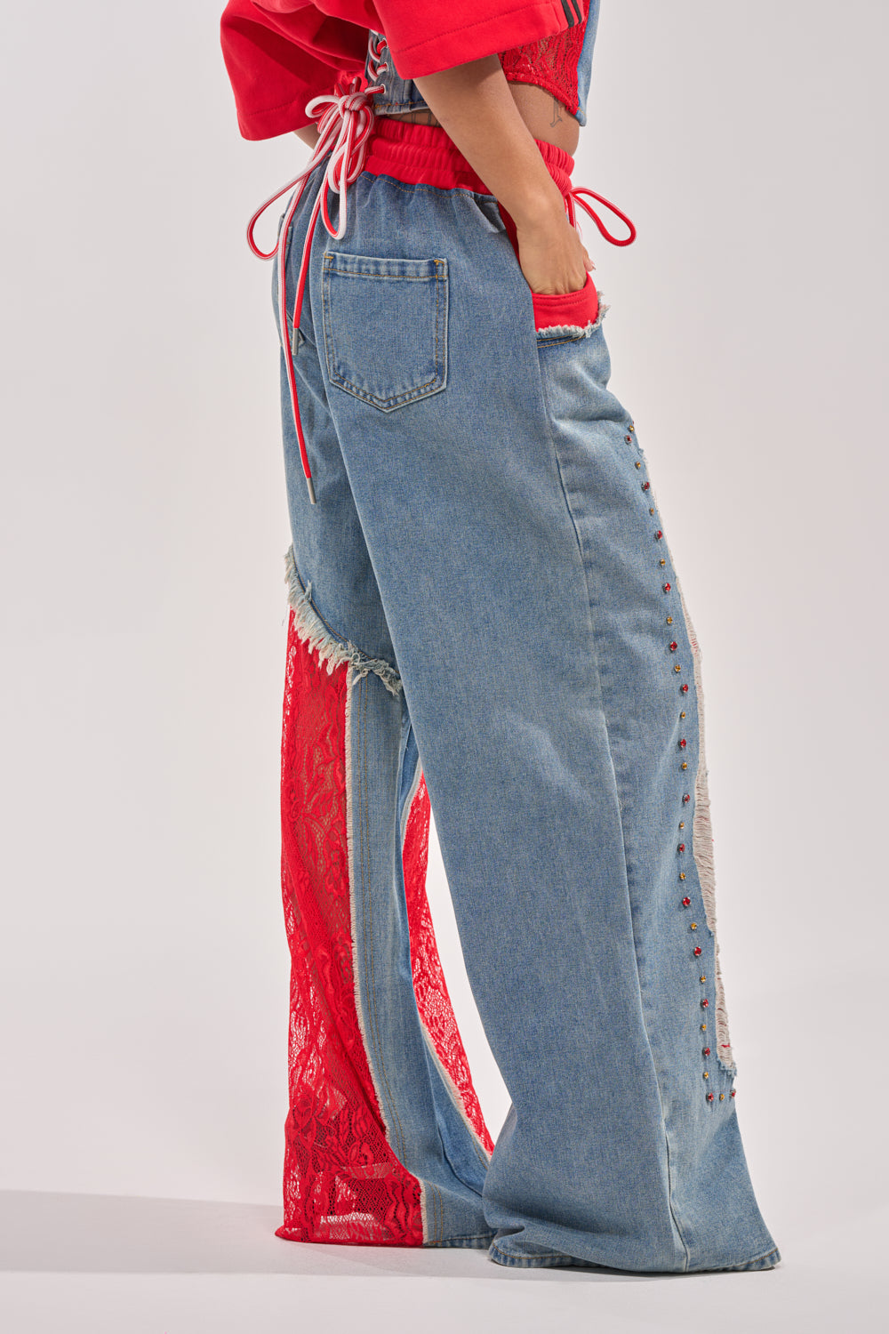 Wearing the GAME TIME JOGGER—patchwork denim joggers with red lace inserts and studded seams—paired with a red drawstring top, this person creates a bold statement streetwear look, standing confidently with one hand in their pocket. 2