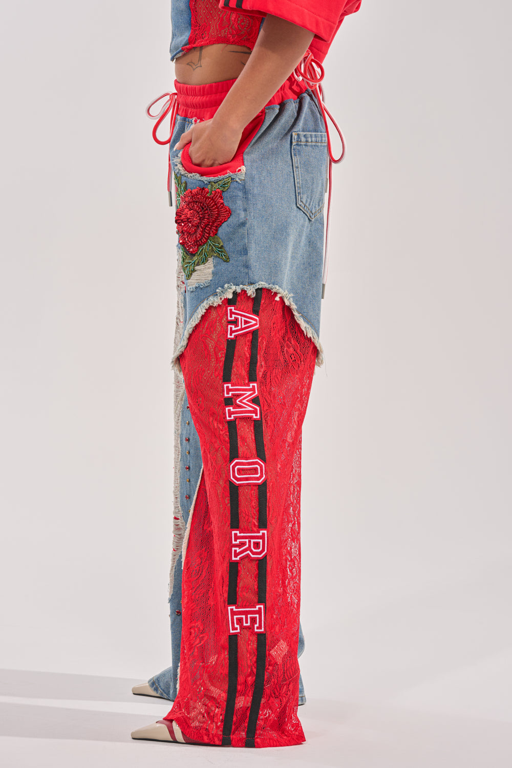 A person stands in profile, wearing the GAME TIME JOGGER: patchwork denim joggers with shorts featuring a red rose patch over red lace pant legs and "AMORE" printed vertically in white on black-striped lace. 4
