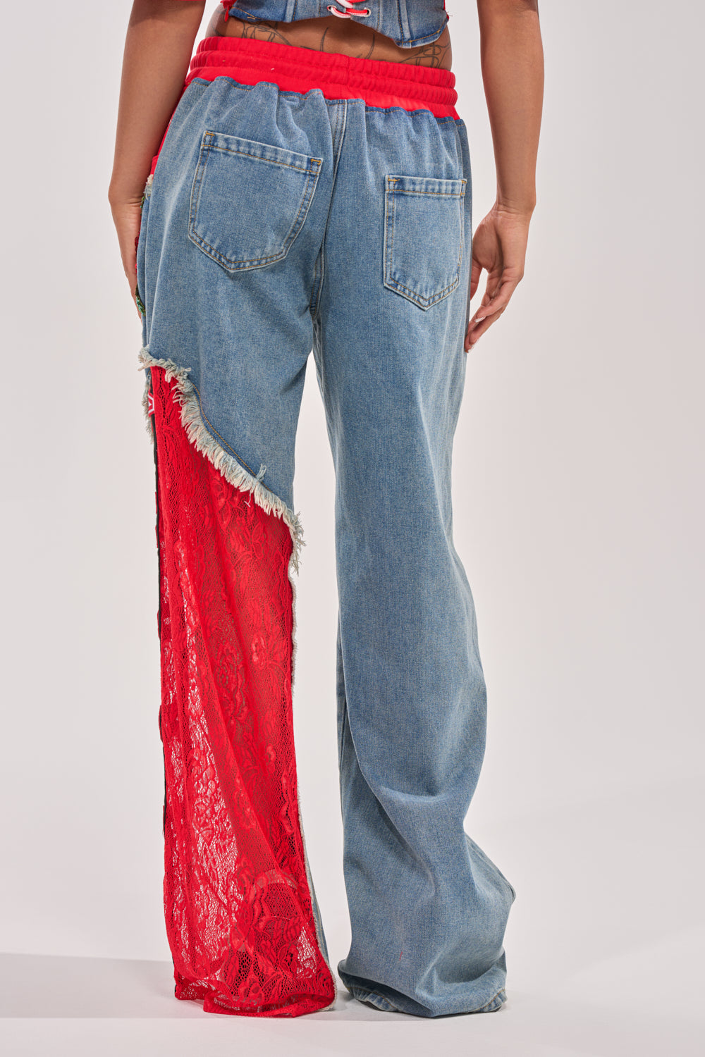A person wearing the GAME TIME JOGGER: blue denim jeans with two back pockets; the left leg features a red lace and fringe panel, inspired by red lace joggers, contrasting with the solid denim right leg. 5
