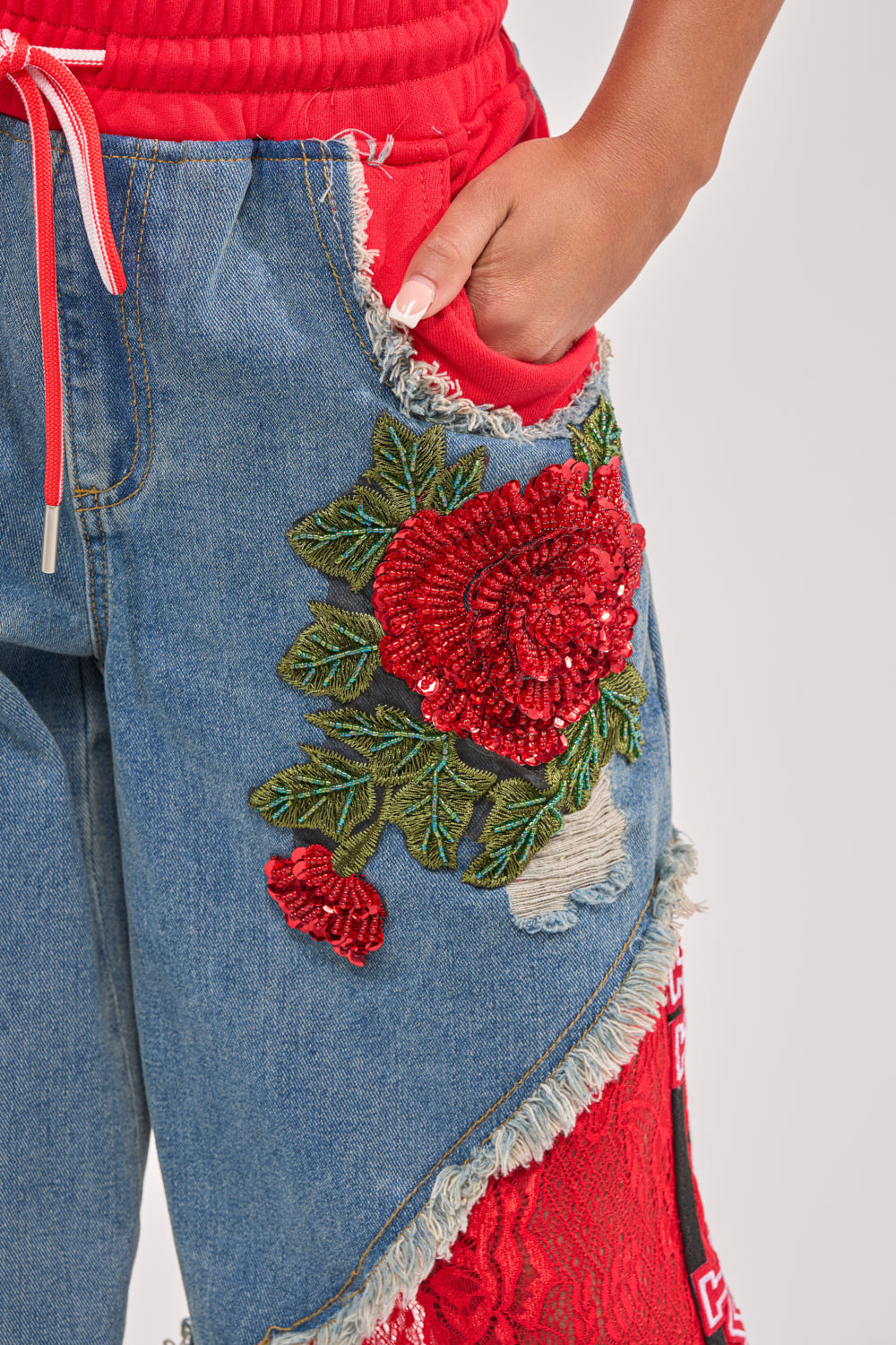 Close-up of a person wearing the GAME TIME JOGGER, featuring patchwork denim, frayed edges, red lace panels, and bold red rose embroidery. Their hand is in the pocket, styled with a red top for standout streetwear lovers. 8
