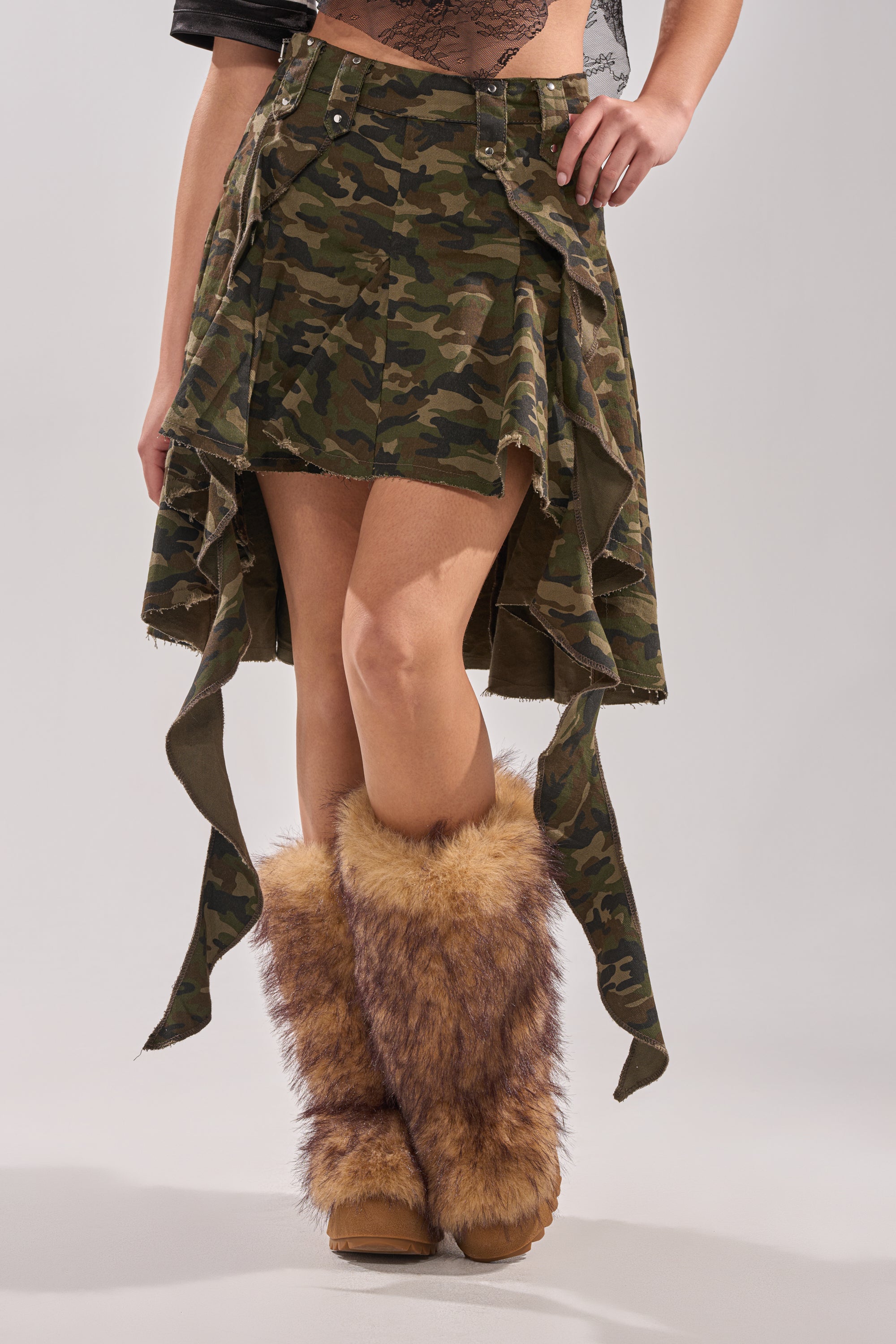 Wearing the IN THE TRENCHES CAMO MINI SKIRT and furry brown leg warmers, a person poses stylishly against a plain light background in this distinctive look. 1