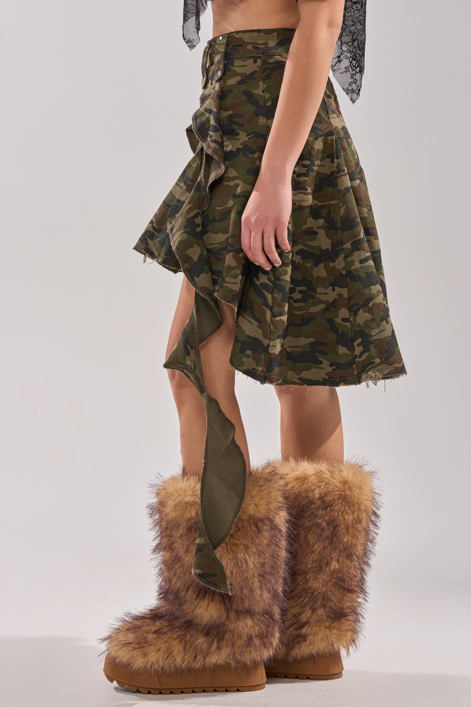 Wearing the IN THE TRENCHES CAMO MINI SKIRT with ruffled details and large brown furry boots, a person stands against a plain light gray background, showing only their lower torso and legs. 4