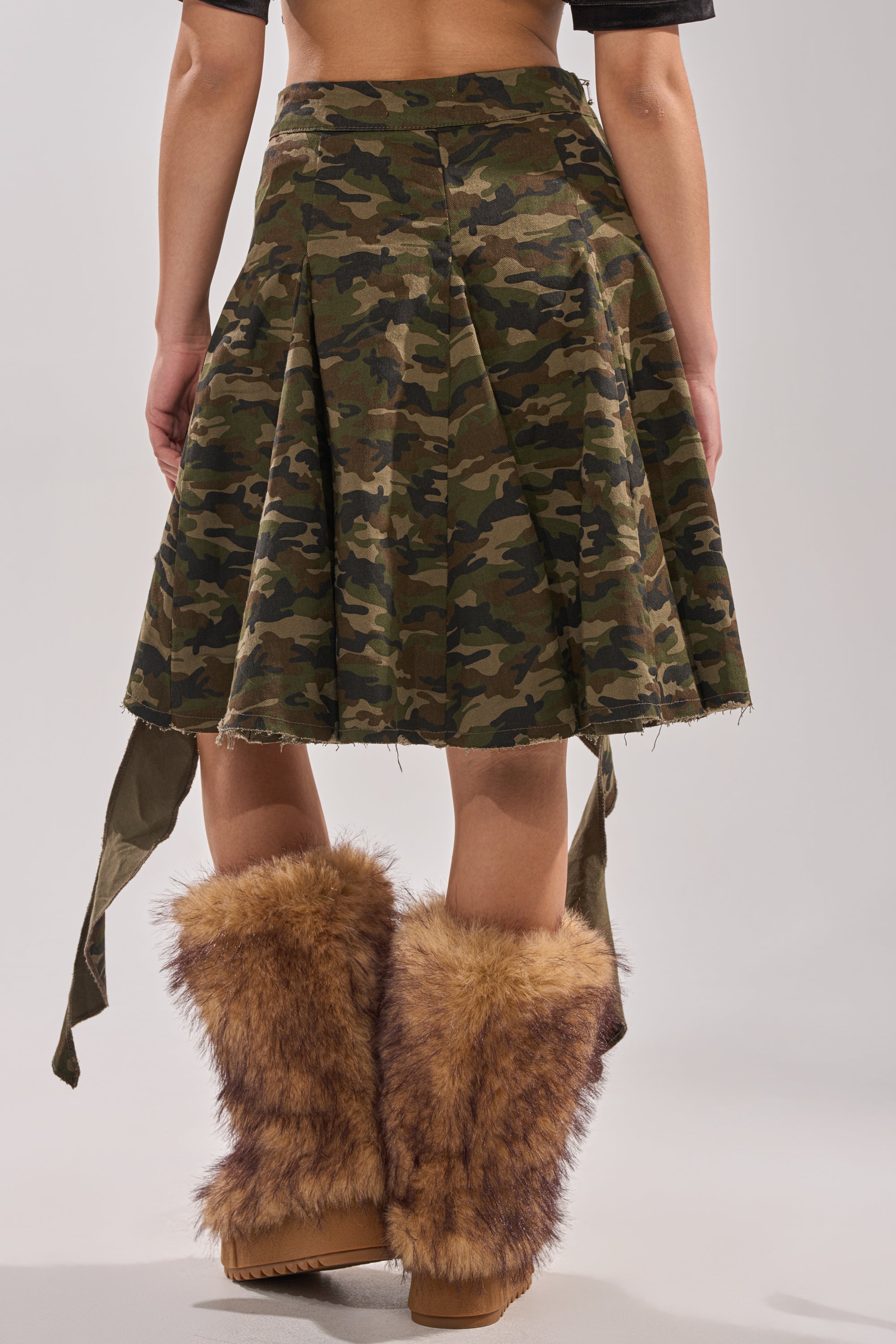 A person wearing the IN THE TRENCHES CAMO MINI SKIRT—a pleated, utility-style, camouflage-print mini skirt—with brown furry boots, shown from the back against a plain light background. 5
