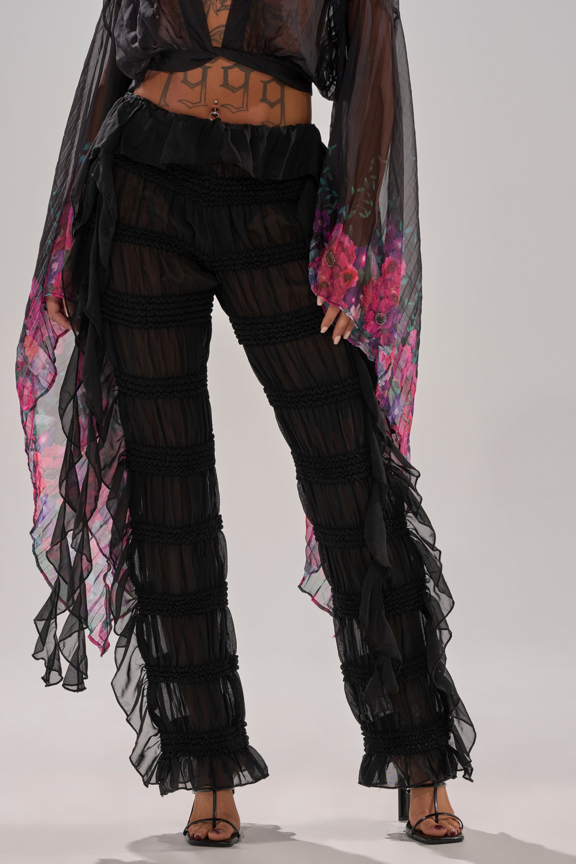 A person wears sheer mesh DANCING QUEEN RUFFLE PANT, black open-toe heels, and a flowy sheer black top with pink and purple floral patterns, the high-rise fit revealing a torso tattoo.