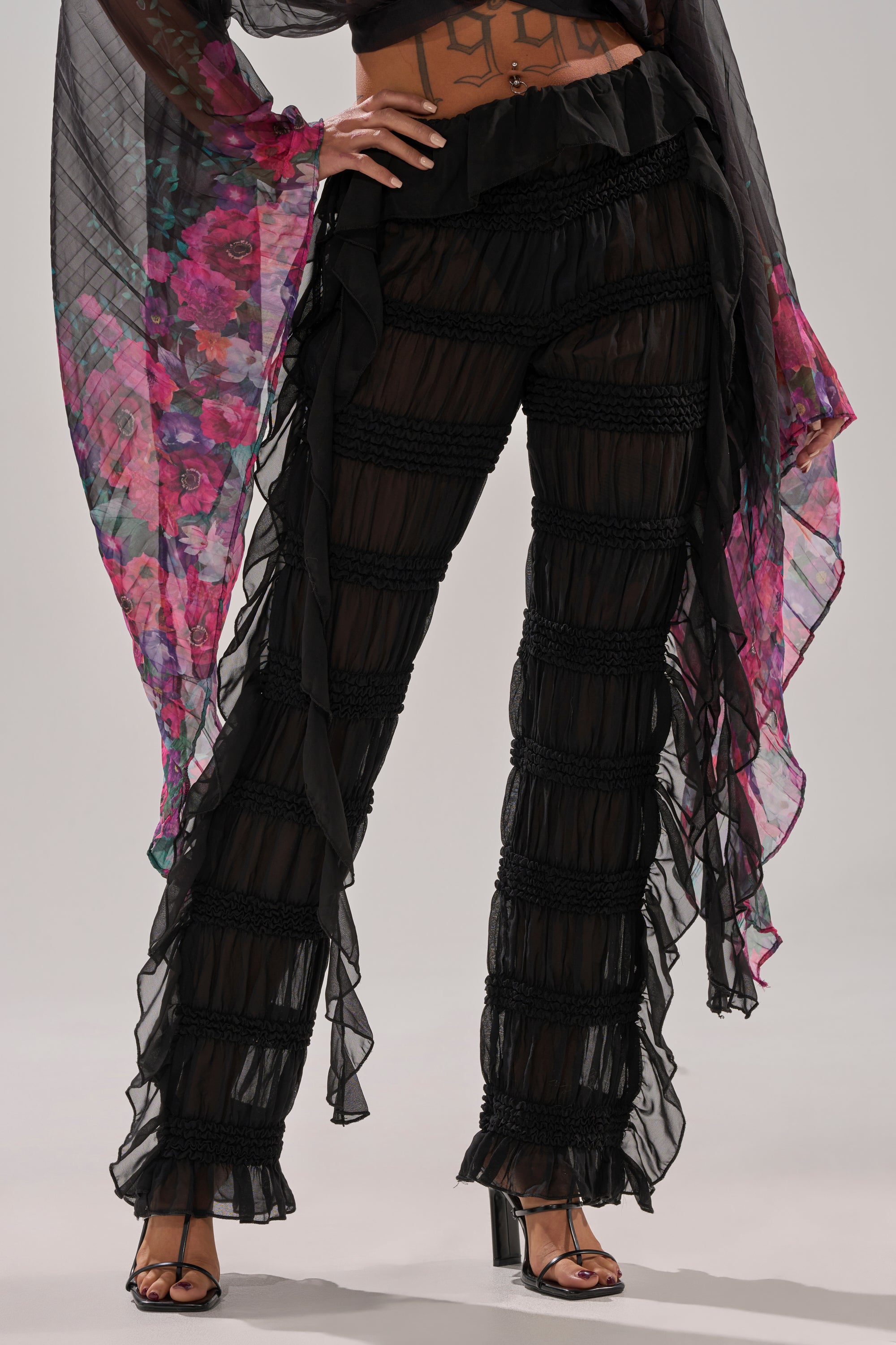 A person wears the DANCING QUEEN RUFFLE PANT, featuring a high-rise fit, sheer black fabric, and horizontal ruched bands, styled with open-toe heels. A flowing floral sheer garment in pink and purple tones appears in the background.