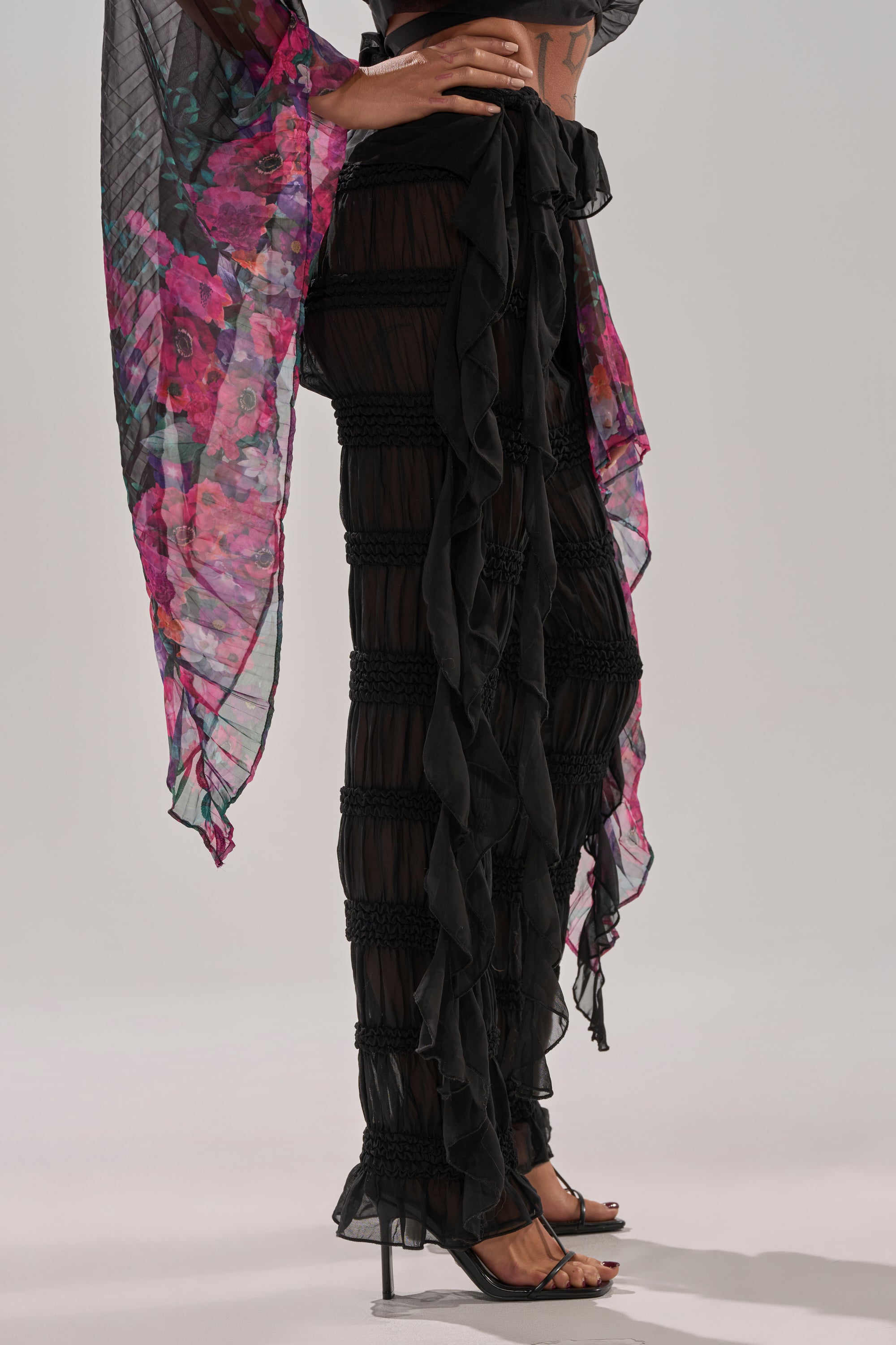 A person wears flowy, sheer DANCING QUEEN RUFFLE PANT with ruffle details and open-toe heels. Their top features translucent pink and purple floral sleeves. The chest-down photo highlights the stylish pants.