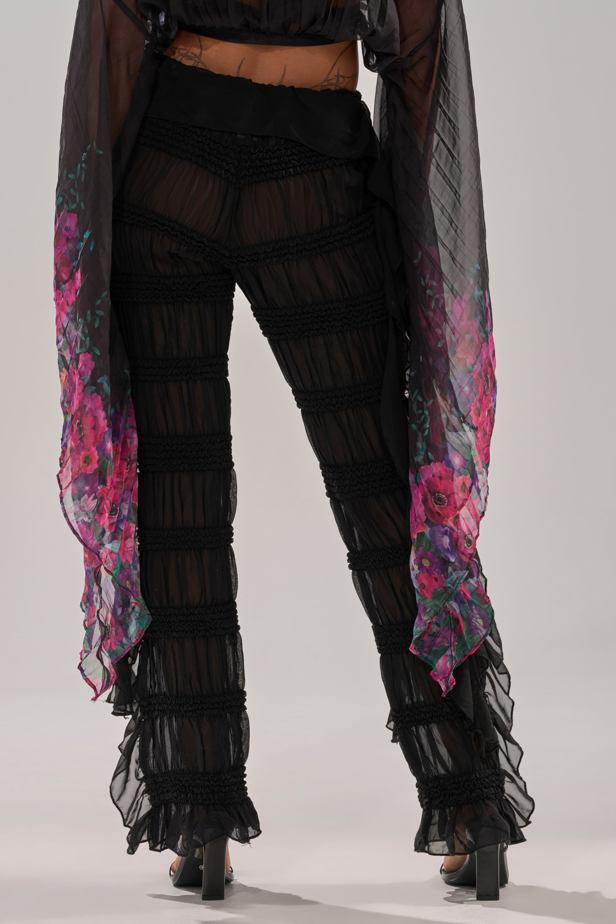 Wearing the DANCING QUEEN RUFFLE PANT—sheer mesh, high-rise fit—paired with a matching sheer black top featuring pink and purple floral details and high-heeled sandals, she perfectly channels Dancing Queen vibes.