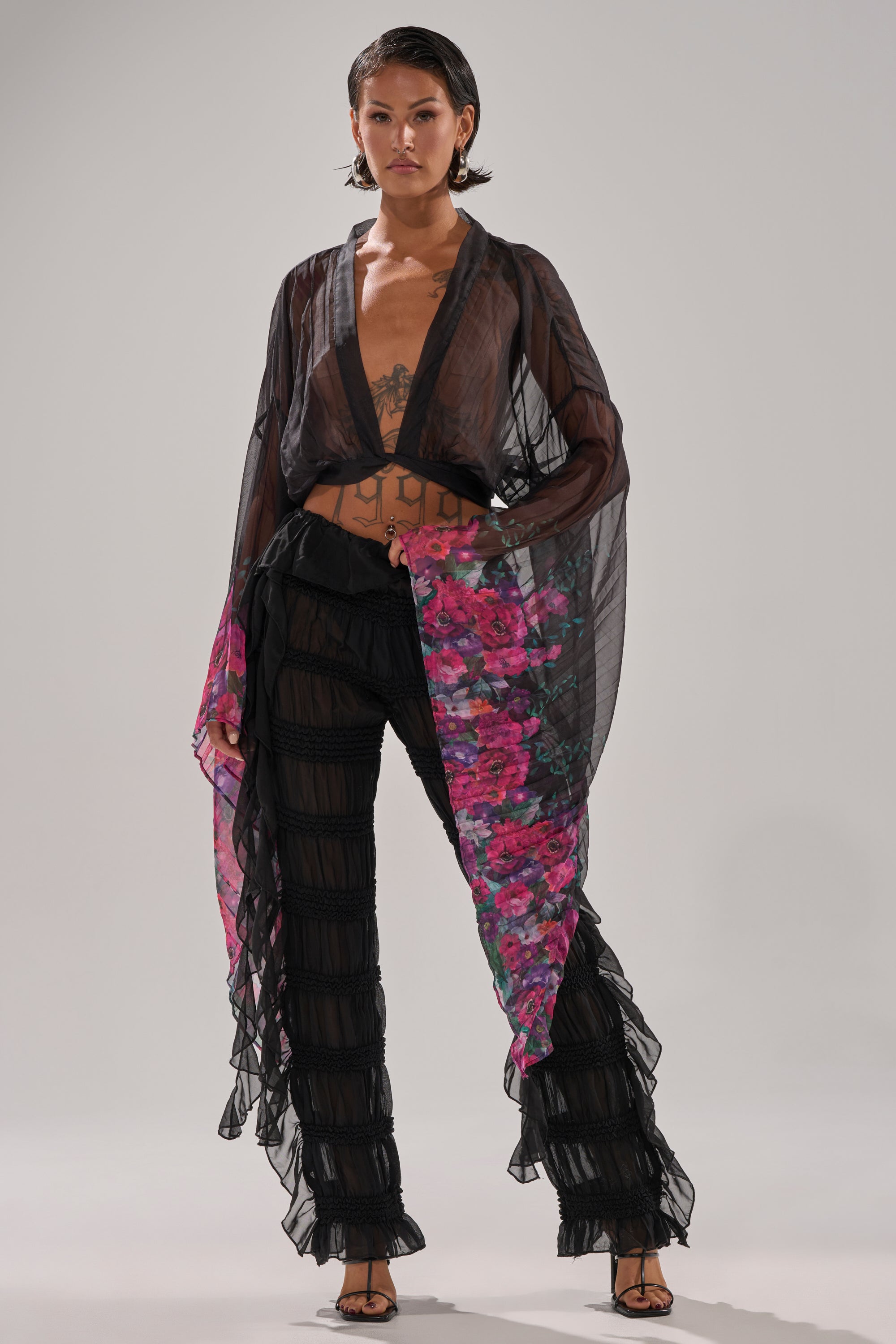 A woman in a sheer black blouse with a deep neckline and pink sleeve accents pairs the DANCING QUEEN RUFFLE PANT—high-rise sheer mesh pants—with black heels, posing confidently against a light background.