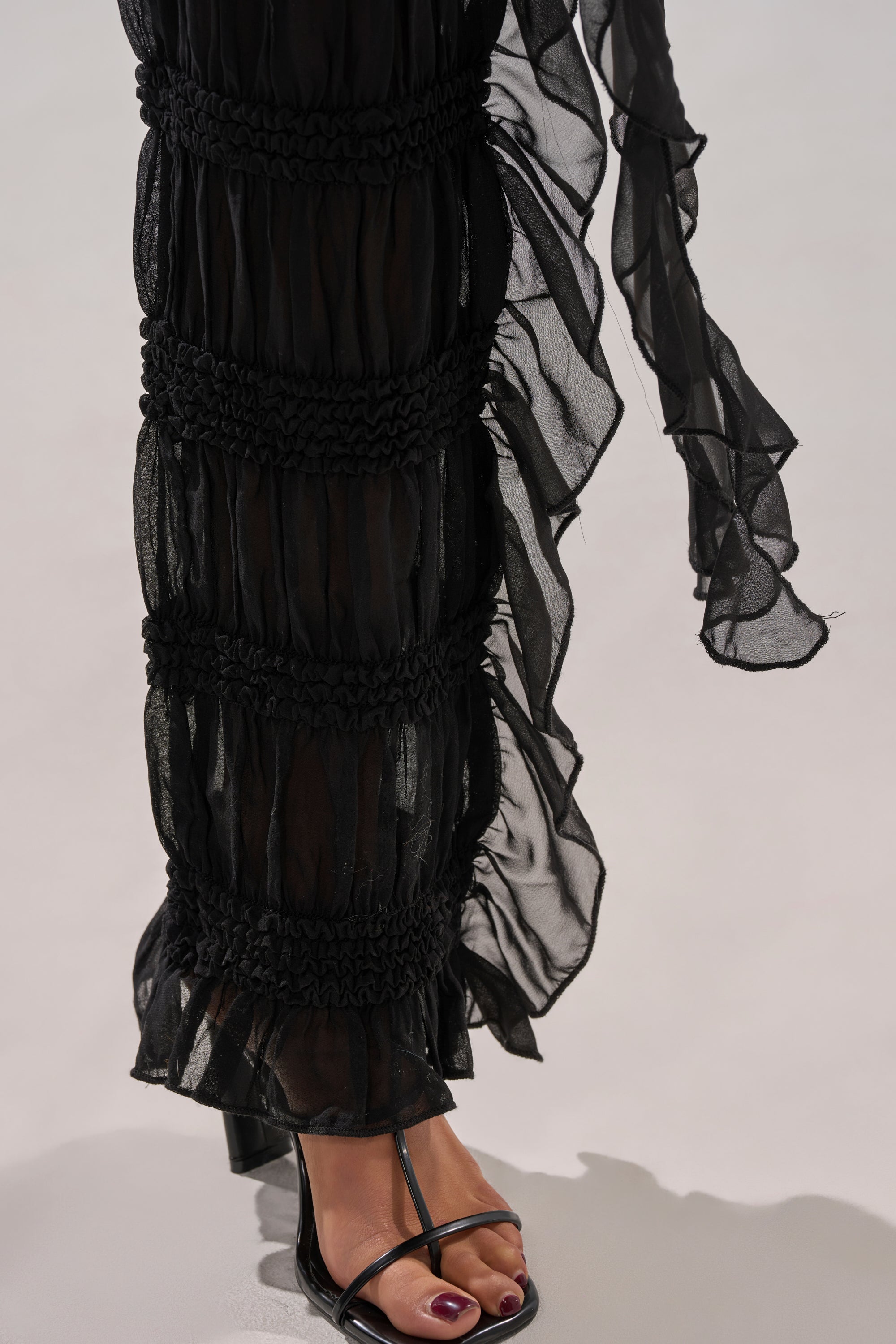 A close-up of a person wearing the DANCING QUEEN RUFFLE PANT—sheer black mesh, high-rise fit—paired with black strappy high-heeled sandals and dark red toenails. The background is plain and light-colored.