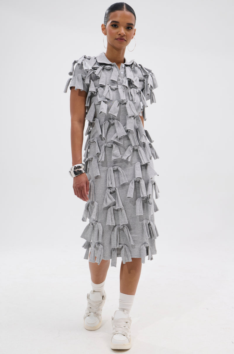 A woman wears the TIED DOWN KNOT MIDI DRESS in light gray, featuring bow-like knot details, styled with white sneakers and statement bracelets, set against a plain white background.