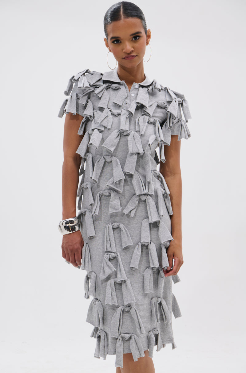 A woman stands against a white background wearing the TIED DOWN KNOT MIDI DRESS, a grey midi dress with hand-tied knots. Her hair is pulled back, she wears hoop earrings, holds one arm at her side, and has a neutral expression.
