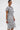 A woman stands against a white background wearing the TIED DOWN KNOT MIDI DRESS, a knee-length gray dress with short sleeves and hand-tied knots. She accessorizes with hoop earrings, a silver bracelet, and has slicked-back hair.