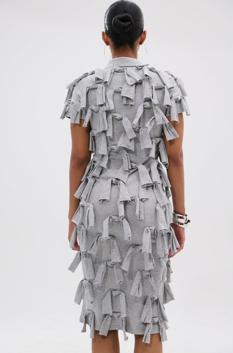 A person faces away from the camera in the TIED DOWN KNOT MIDI DRESS, a fitted, hand-tied knot cotton dress in gray. The knee-length style is styled with a neat bun, hoop earrings, and bracelets for a chic look.