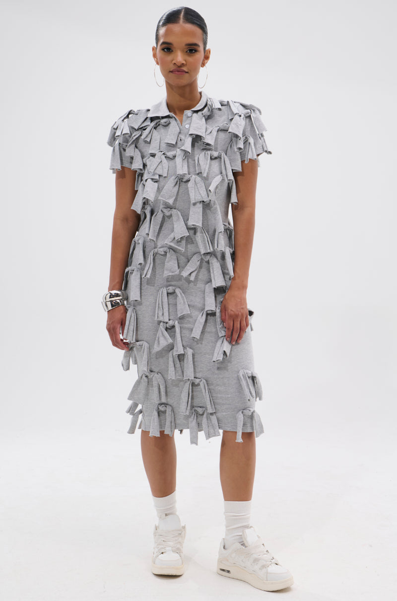A woman poses against a white background wearing the TIED DOWN KNOT MIDI DRESS, a knee-length, short-sleeved cotton dress featuring multiple hand-tied knots. She styles it with white sneakers, socks, and silver bracelets.