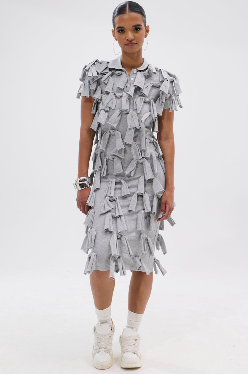 A woman stands against a white background, wearing the TIED DOWN KNOT MIDI DRESS in light gray cotton with hand-tied knots. She pairs it with white sneakers, socks, chunky silver bracelets, and has her hair pulled back.