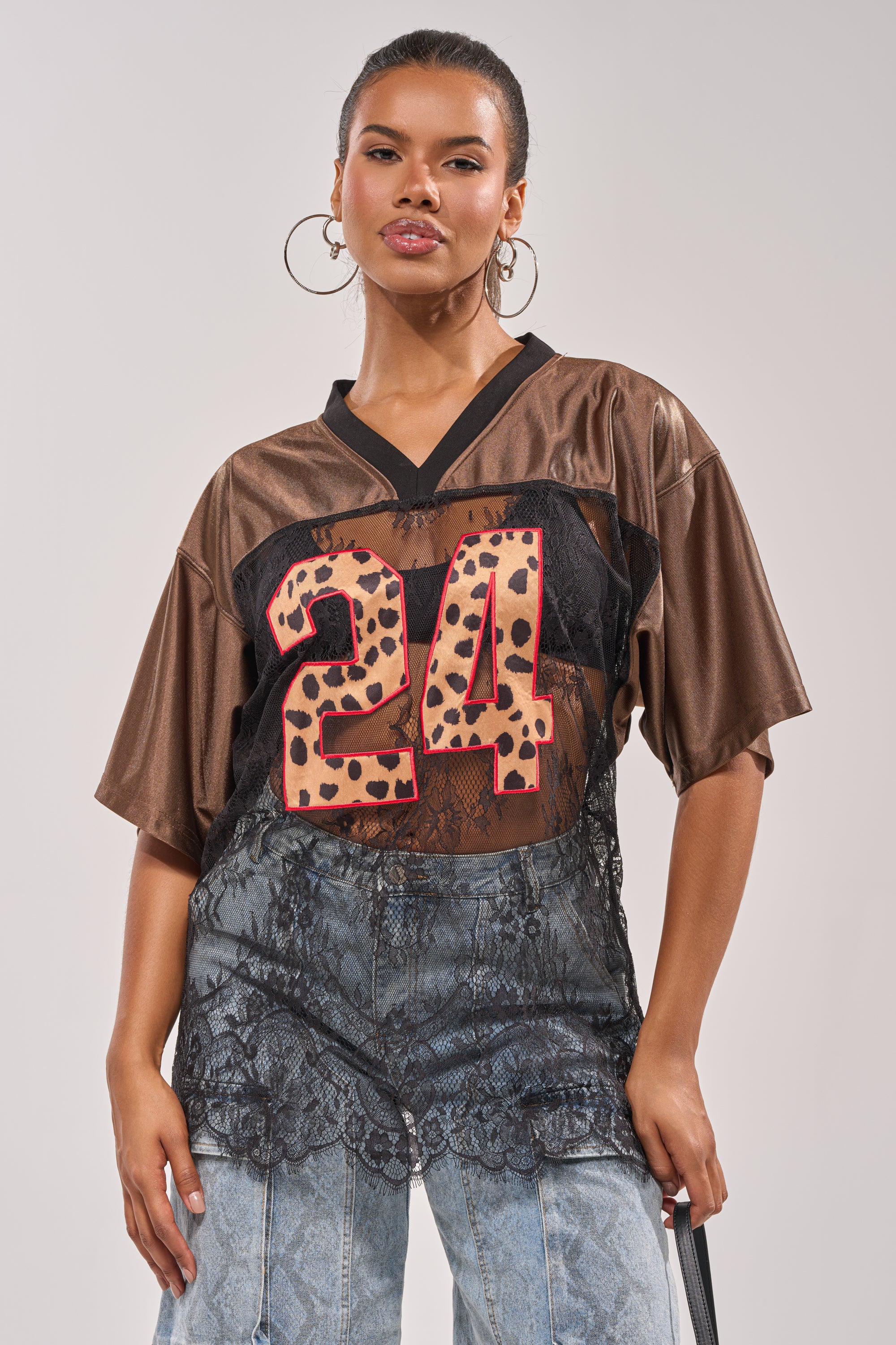 A woman stands confidently against a plain background wearing hoop earrings, the LUCKY 24 JERSEY with a leopard print "24" graphic, and high-waisted blue jeans with sheer lace paneling. 2