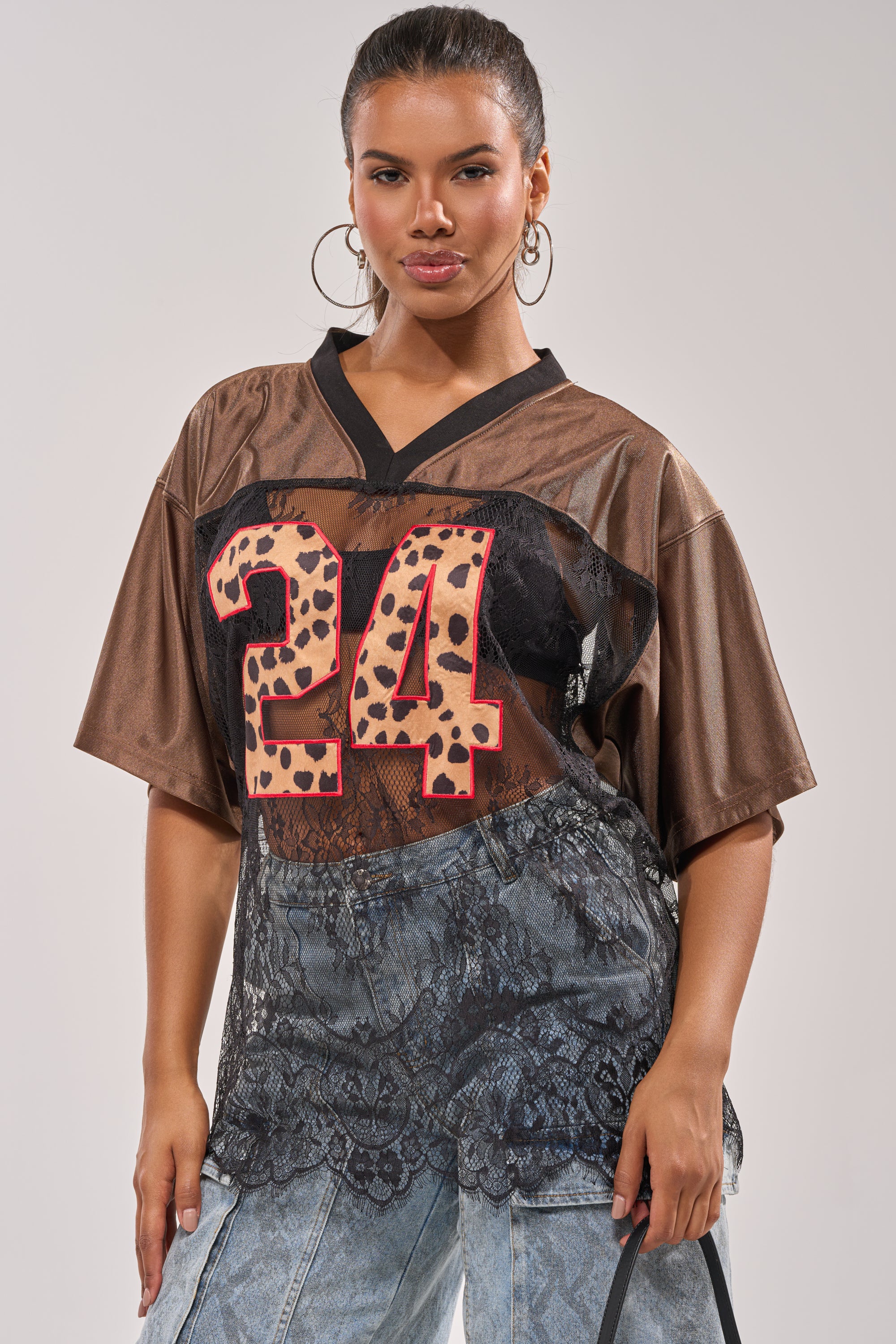 A woman poses confidently against a plain background in the oversized LUCKY 24 JERSEY featuring a leopard print "24" and sheer lace paneling, paired with light-wash denim pants and large hoop earrings for a bold look. 3