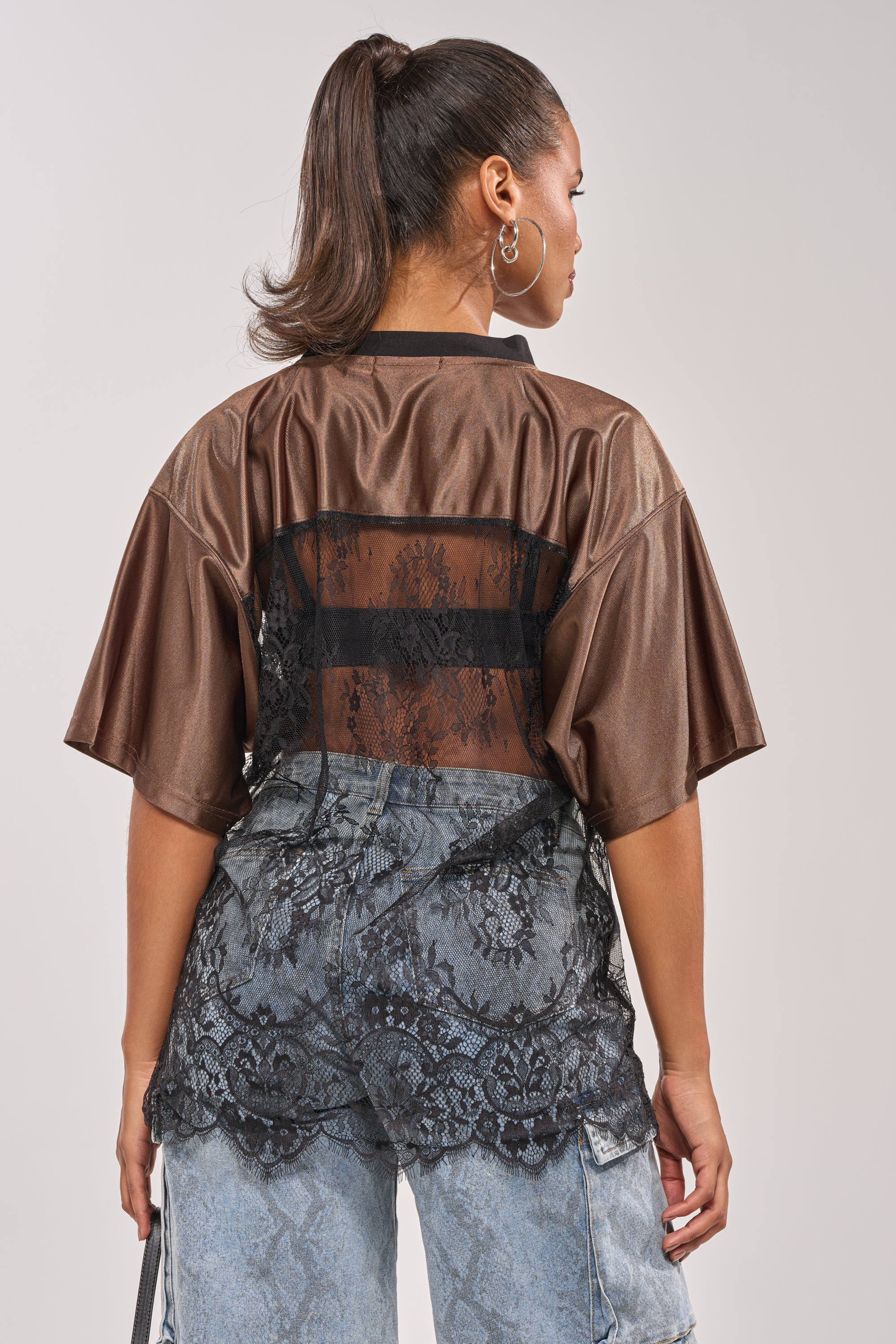 A woman with a high ponytail exudes street-chic style in the LUCKY 24 JERSEY, featuring a brown short-sleeve design with a sheer lace panel back, paired with light-wash distressed jeans with black lace details. She is turned away from the camera. 6