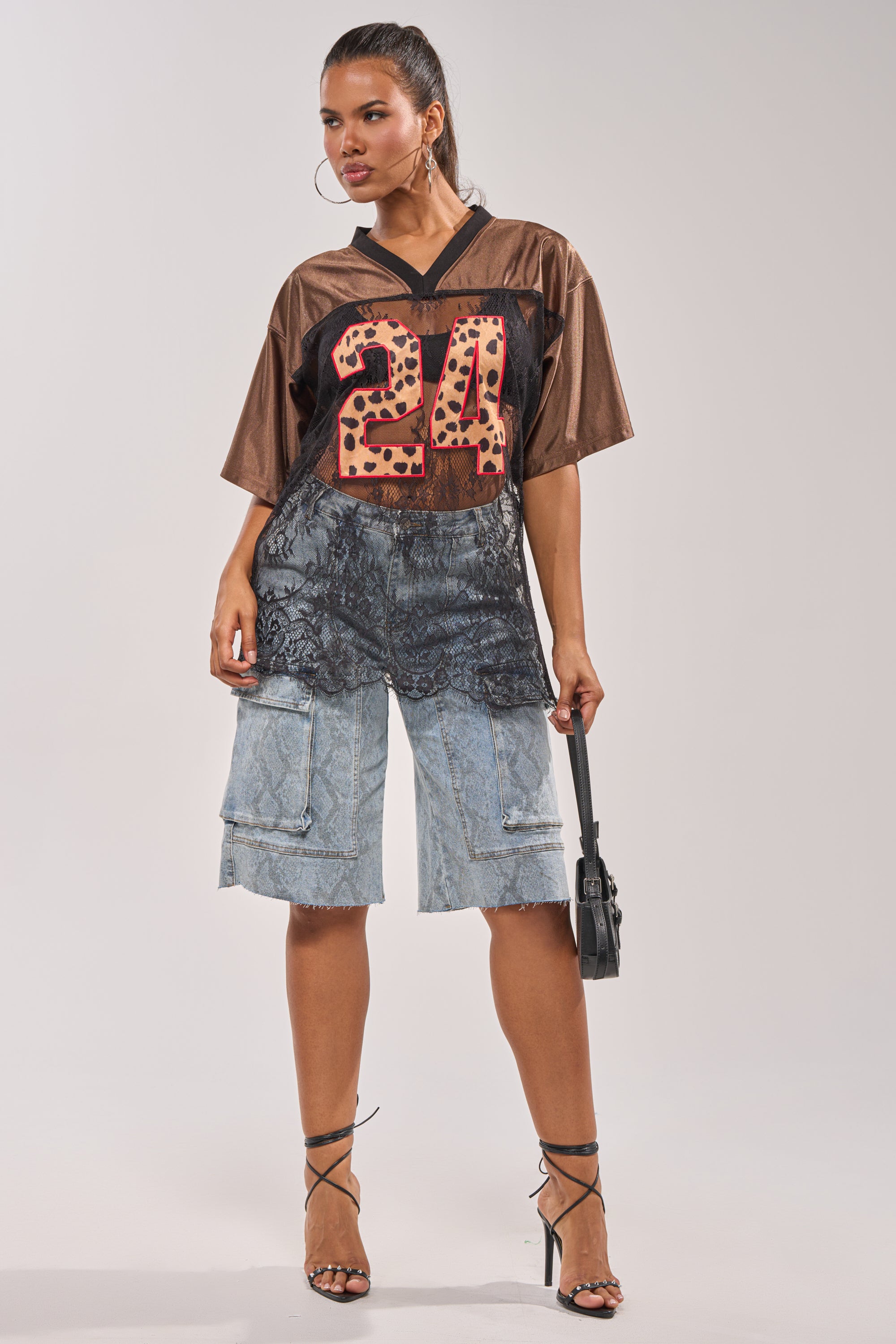 A woman exudes street-chic confidence in the LUCKY 24 JERSEY—a sheer lace panel oversized top with a leopard-print 24—paired with light blue lace-detailed cargo shorts, lace-up heels, hoop earrings, and a small black handbag. 7